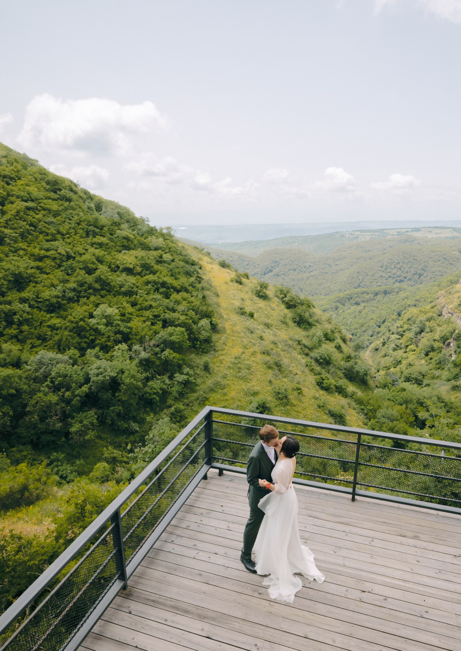 Wedding couple dancing on a scenic mountain overlook deck with panoramic forest views.