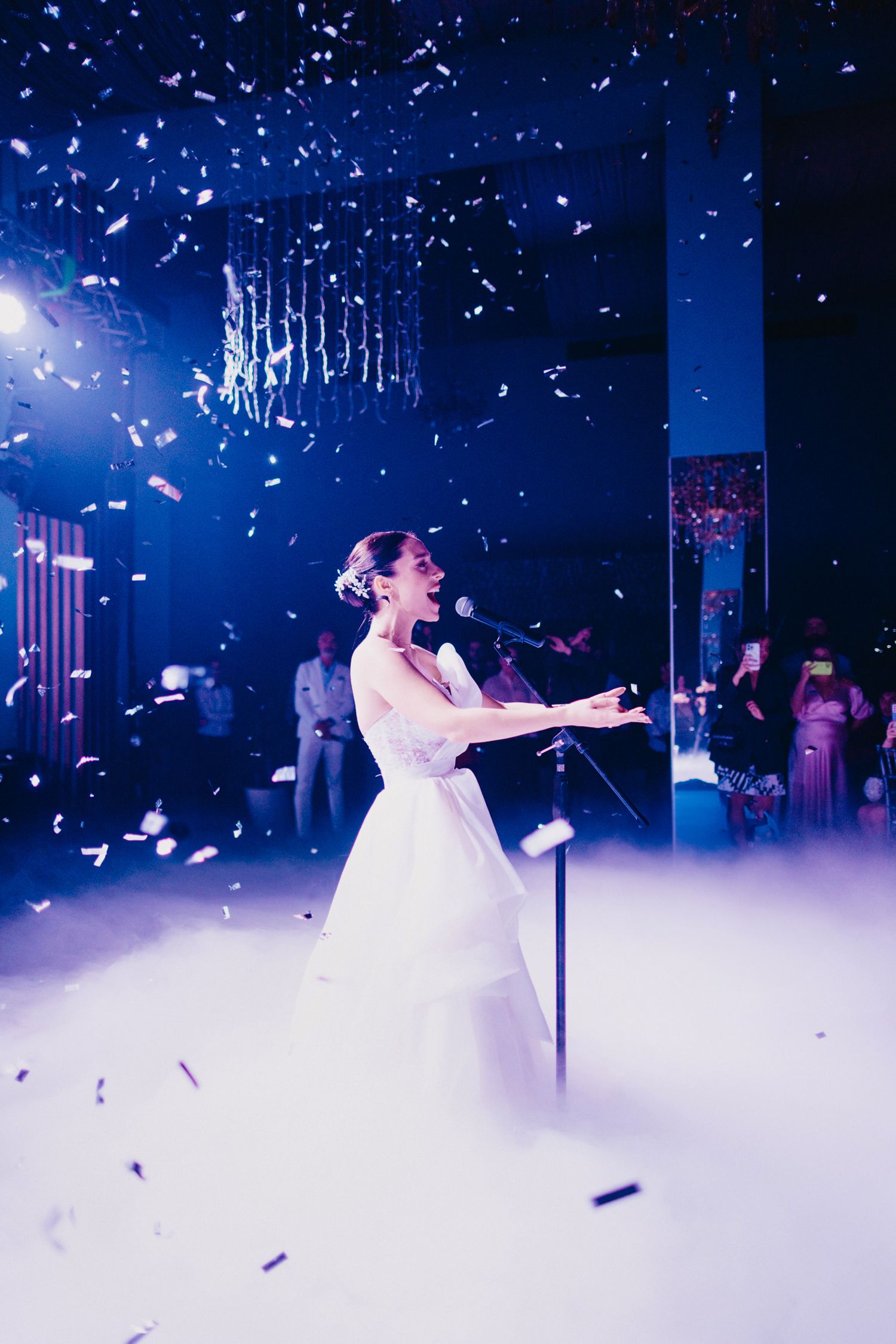 Bride performs on stage surrounded by falling confetti and dramatic blue lighting.