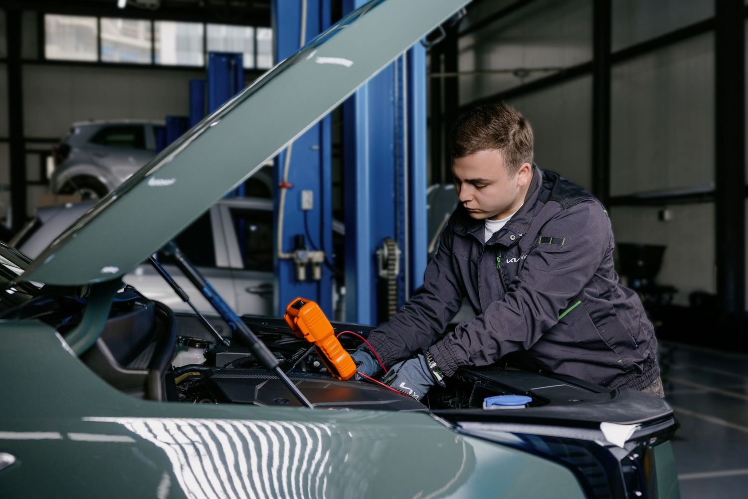 Young mechanic in uniform inspecting car engine under open hood in professional auto repair garage.