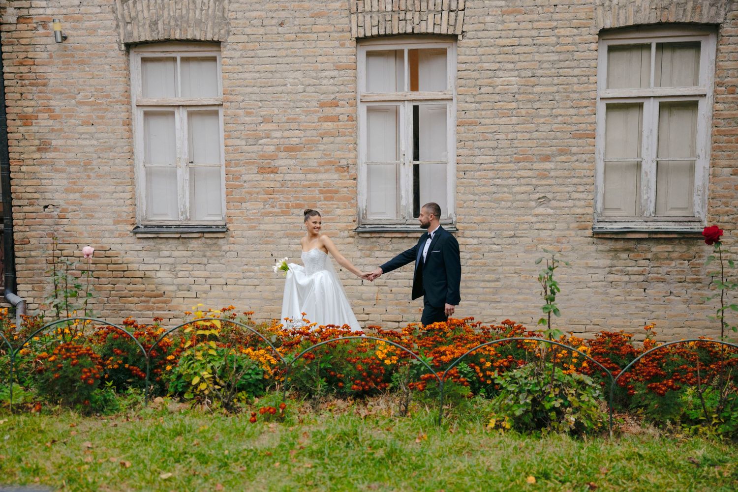 Wedding couple poses by colorful marigold garden against vintage brick building facade.