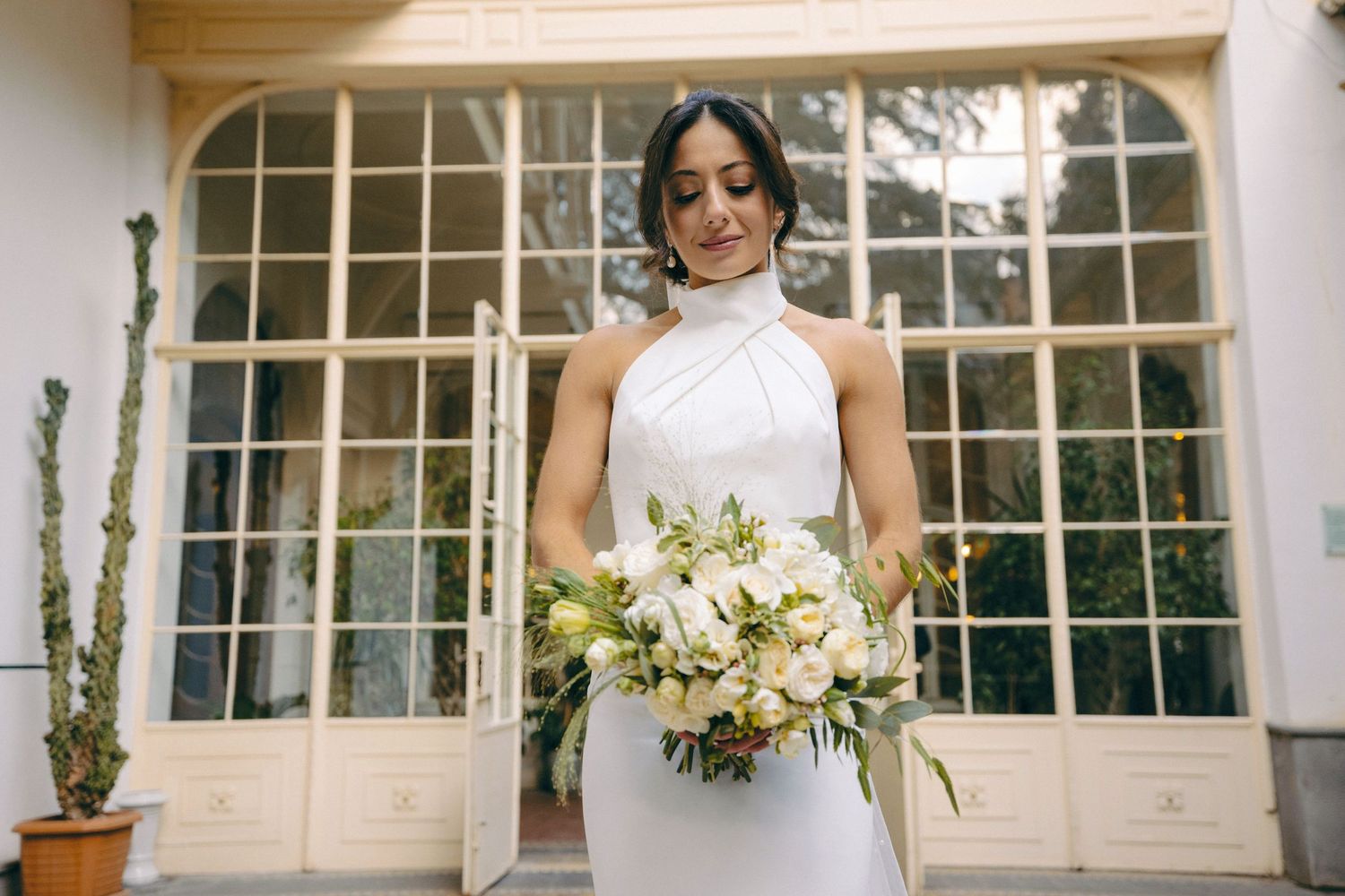Elegant bride poses with bouquet near large windows in classic interior.
