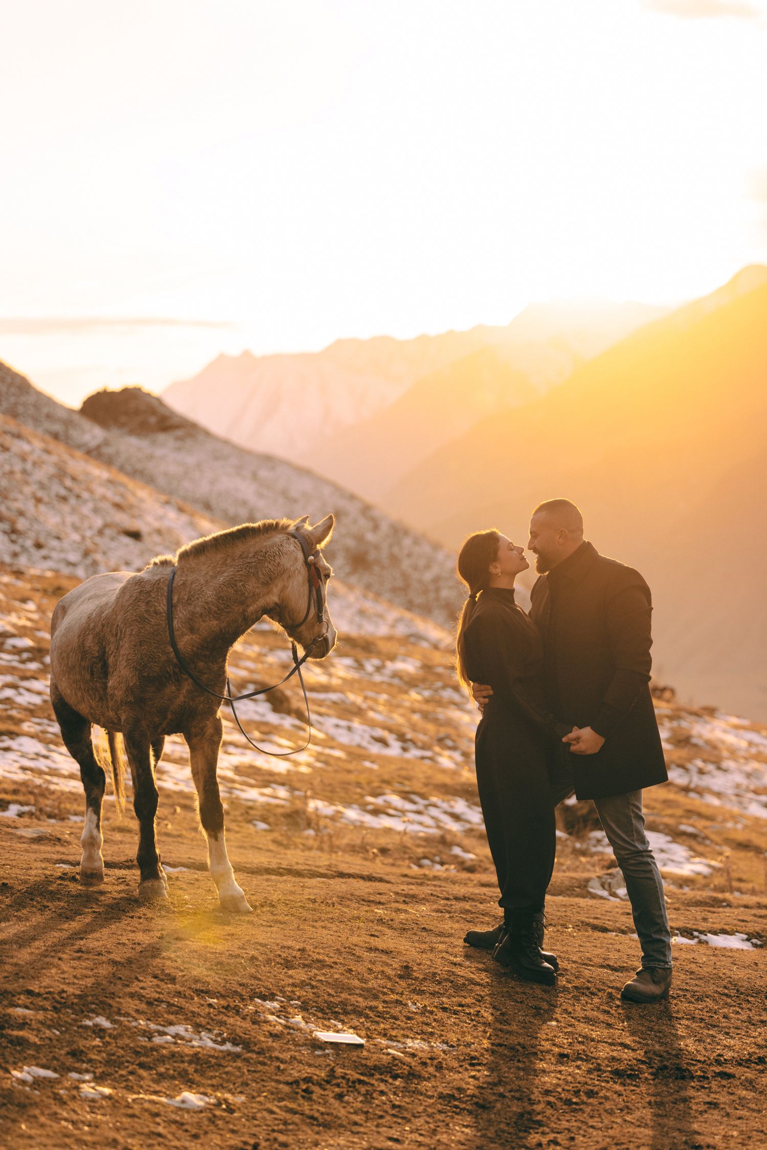Two people share an intimate moment on a snowy mountainside with a horse nearby at sunset.