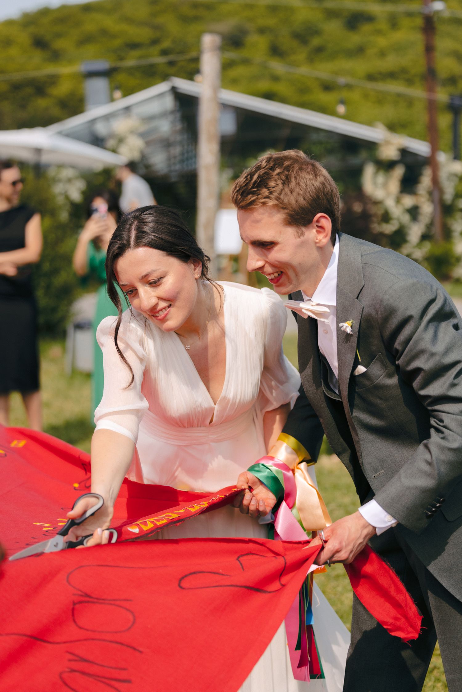 Newlyweds signing a red banner together at their outdoor wedding celebration.