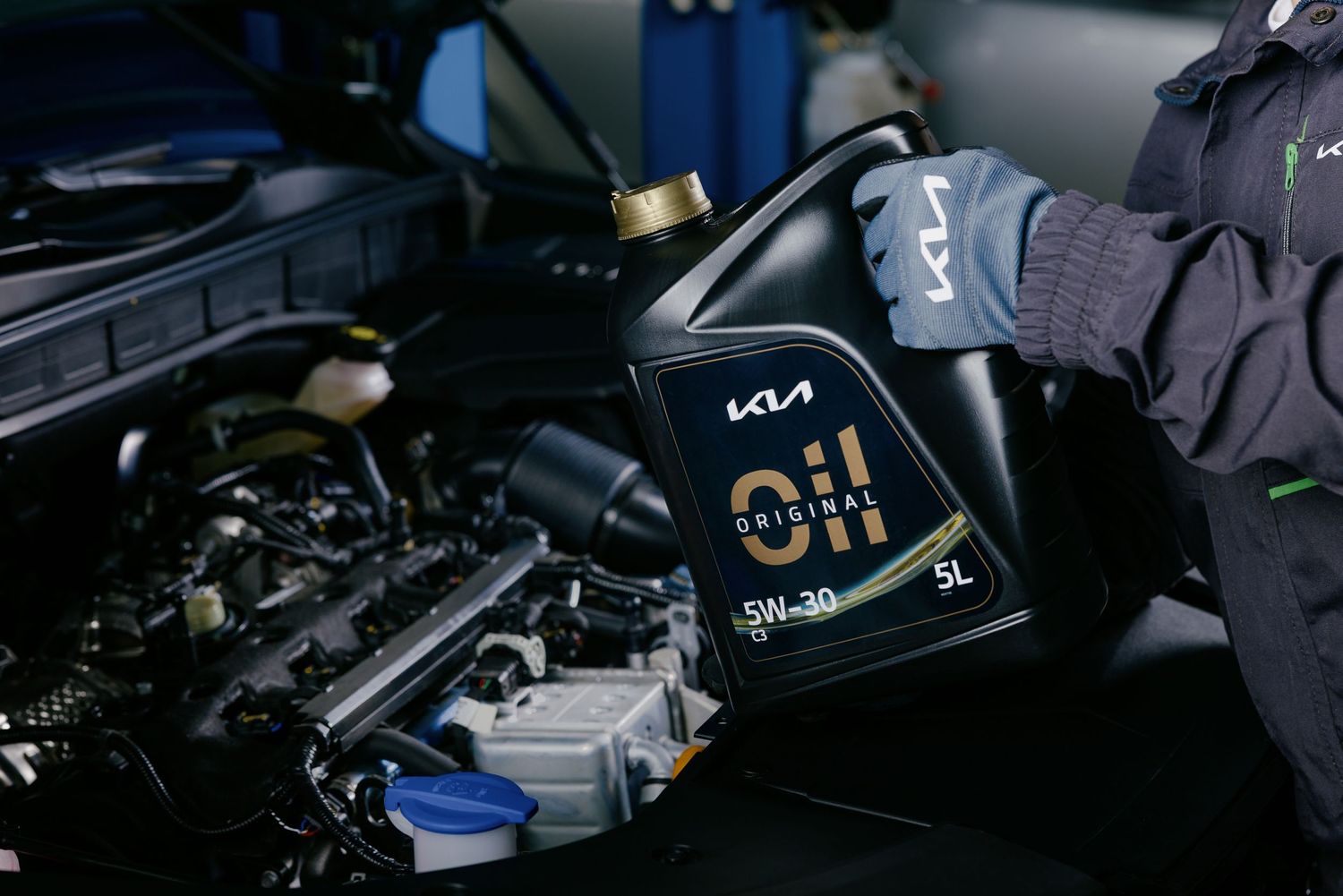 Mechanic holding a 5L Kia Original Oil 5W-30 container over a car engine during a service.