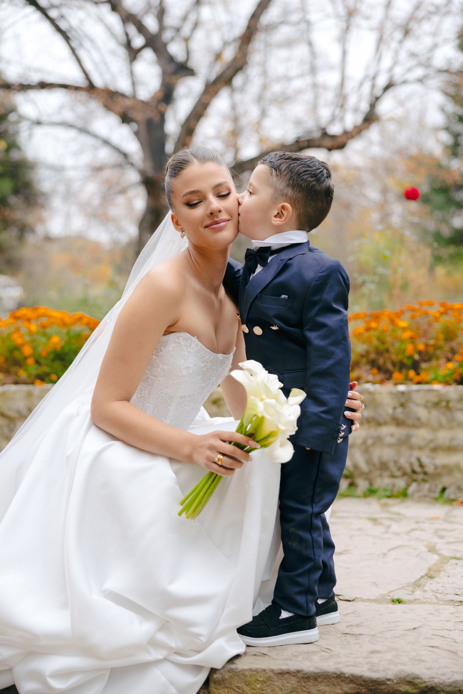 Sweet moment captured between bride in white gown and young ring bearer by garden.