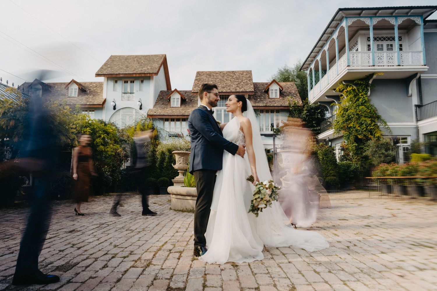 Newlyweds pose for photos at a quaint village venue with cobblestone paths and charming buildings.