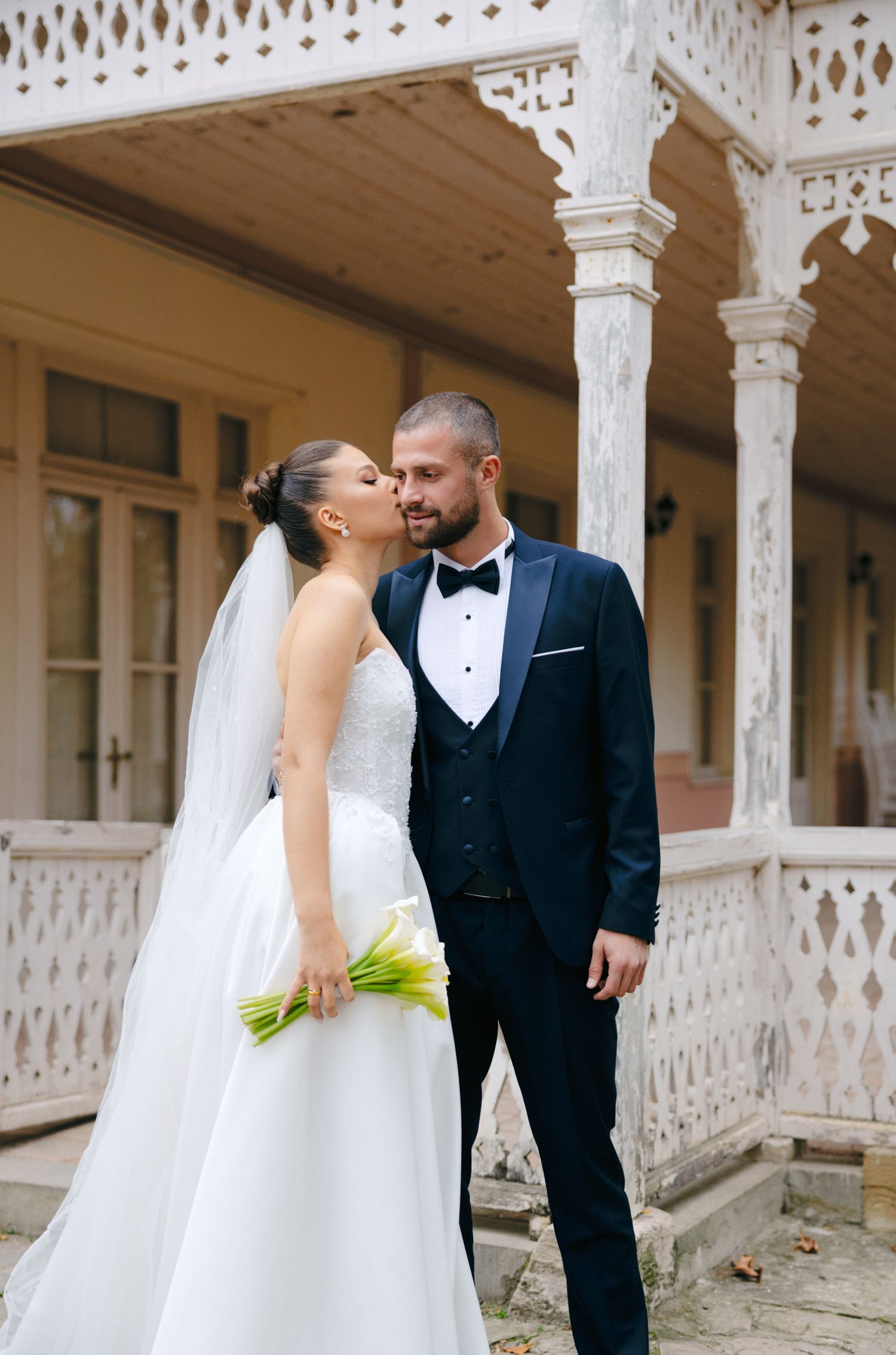 Newlyweds share an intimate moment on a vintage porch with ornate white lattice details.