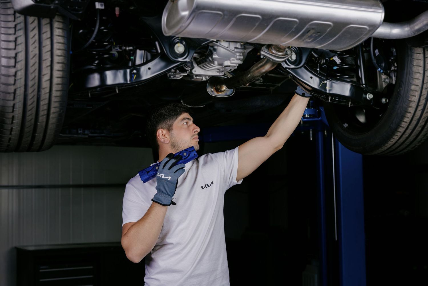 A mechanic in white uniform and blue gloves inspects the undercarriage of a lifted vehicle in an auto repair shop.