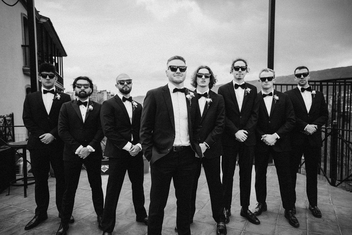 Black and white photo of groomsmen in tuxedos posing outdoors with serious expressions.