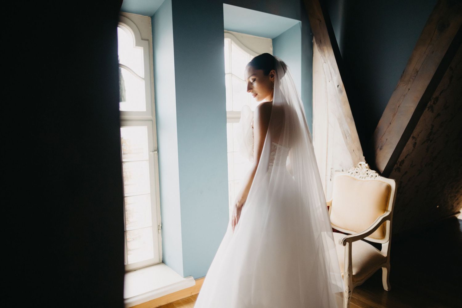Elegant bride in flowing white gown poses near window in moody lighting creating dramatic silhouette.
