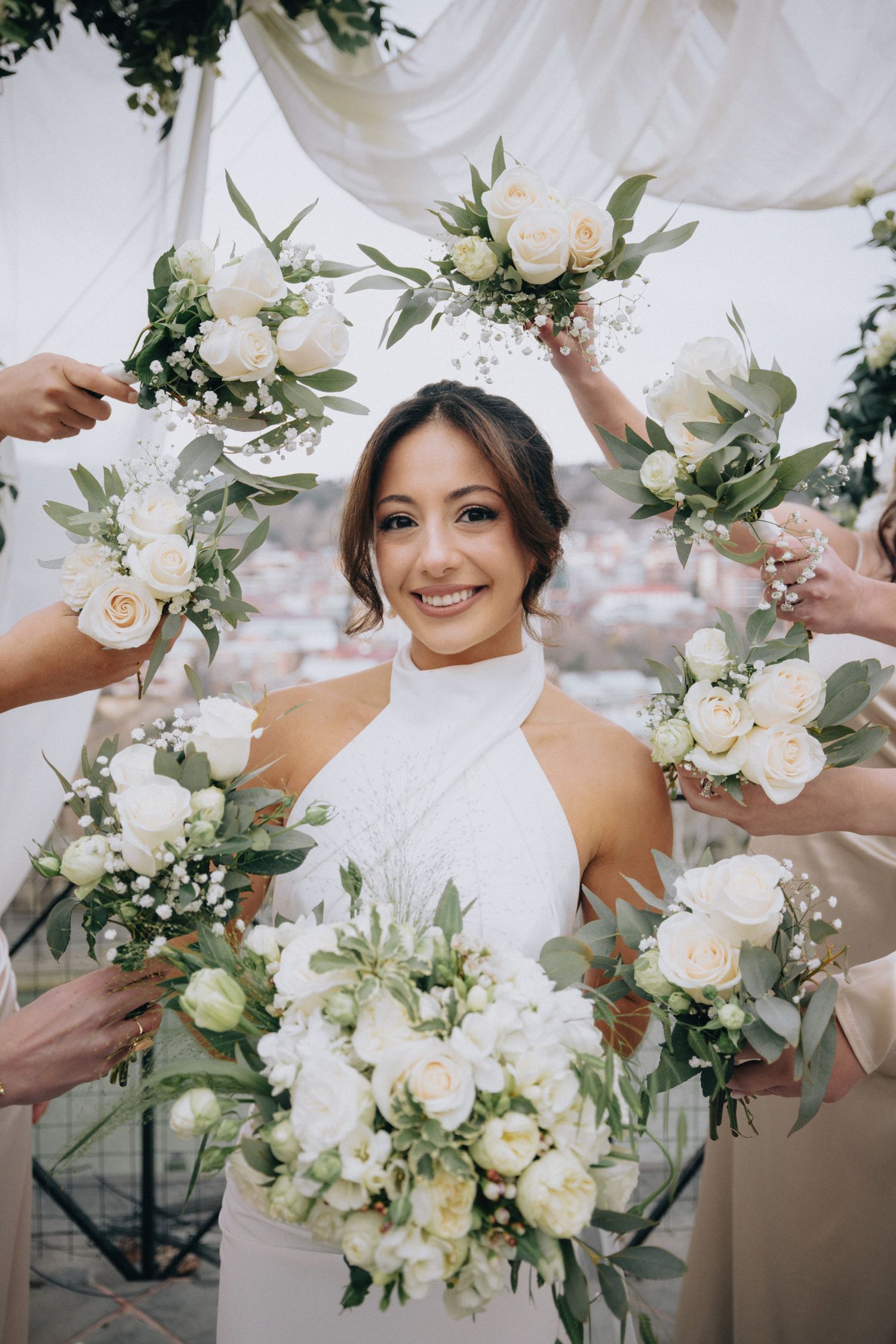 Bride in white halter dress surrounded by raised bouquets of white roses and greenery.
