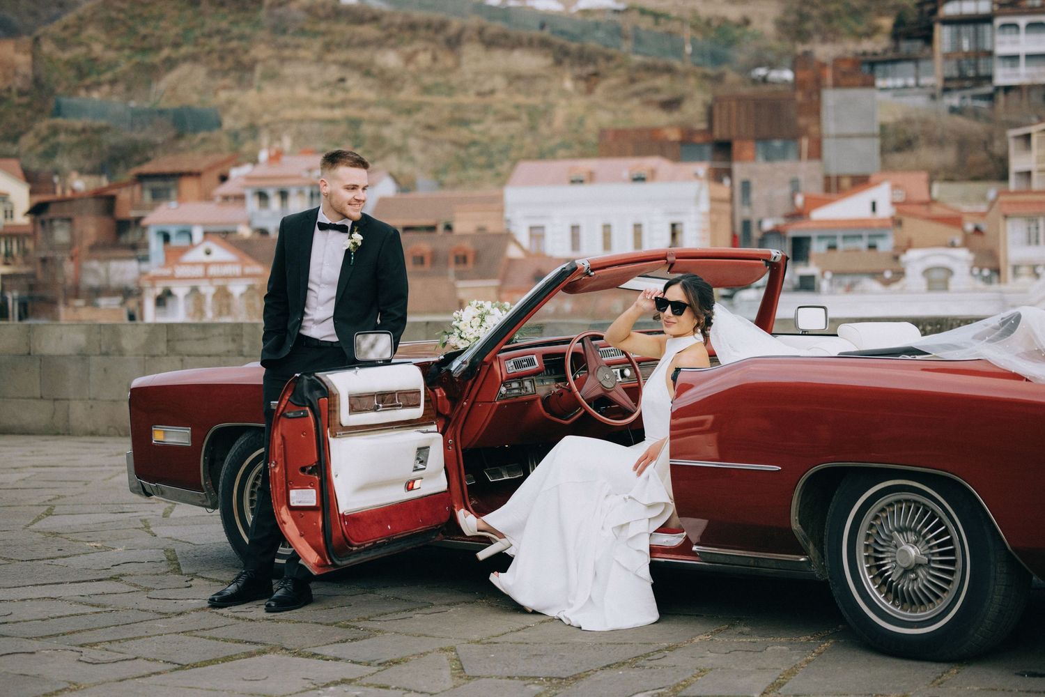 Wedding couple posing with vintage red car against Mediterranean hillside backdrop.