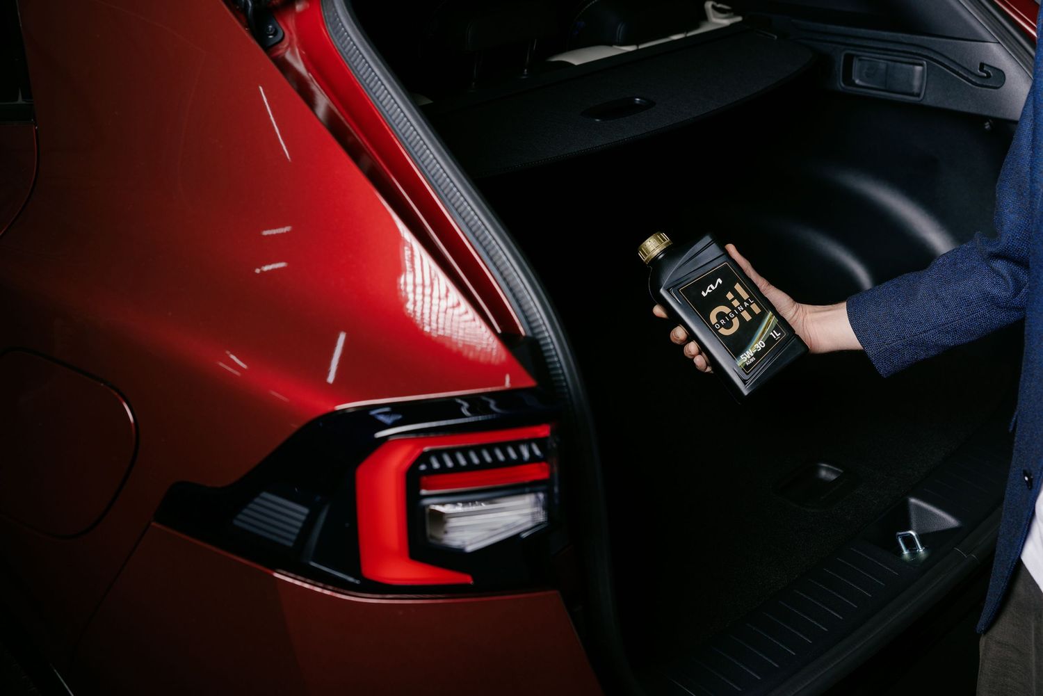 Person placing a dark bottle into the trunk of a red car with illuminated taillights.