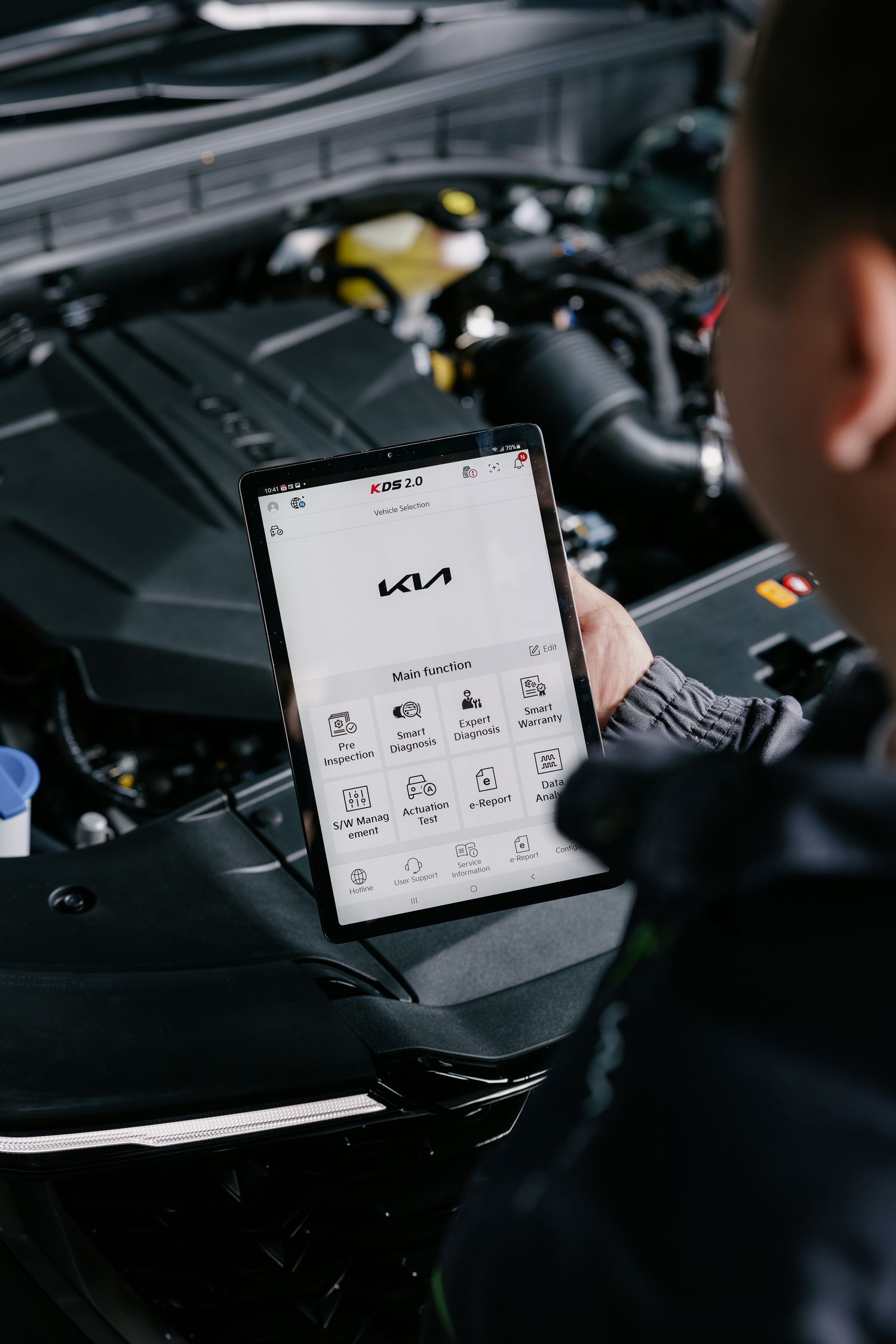 Mechanic using KIA KDS 2.0 diagnostic tablet tool while inspecting a car engine during a service check.