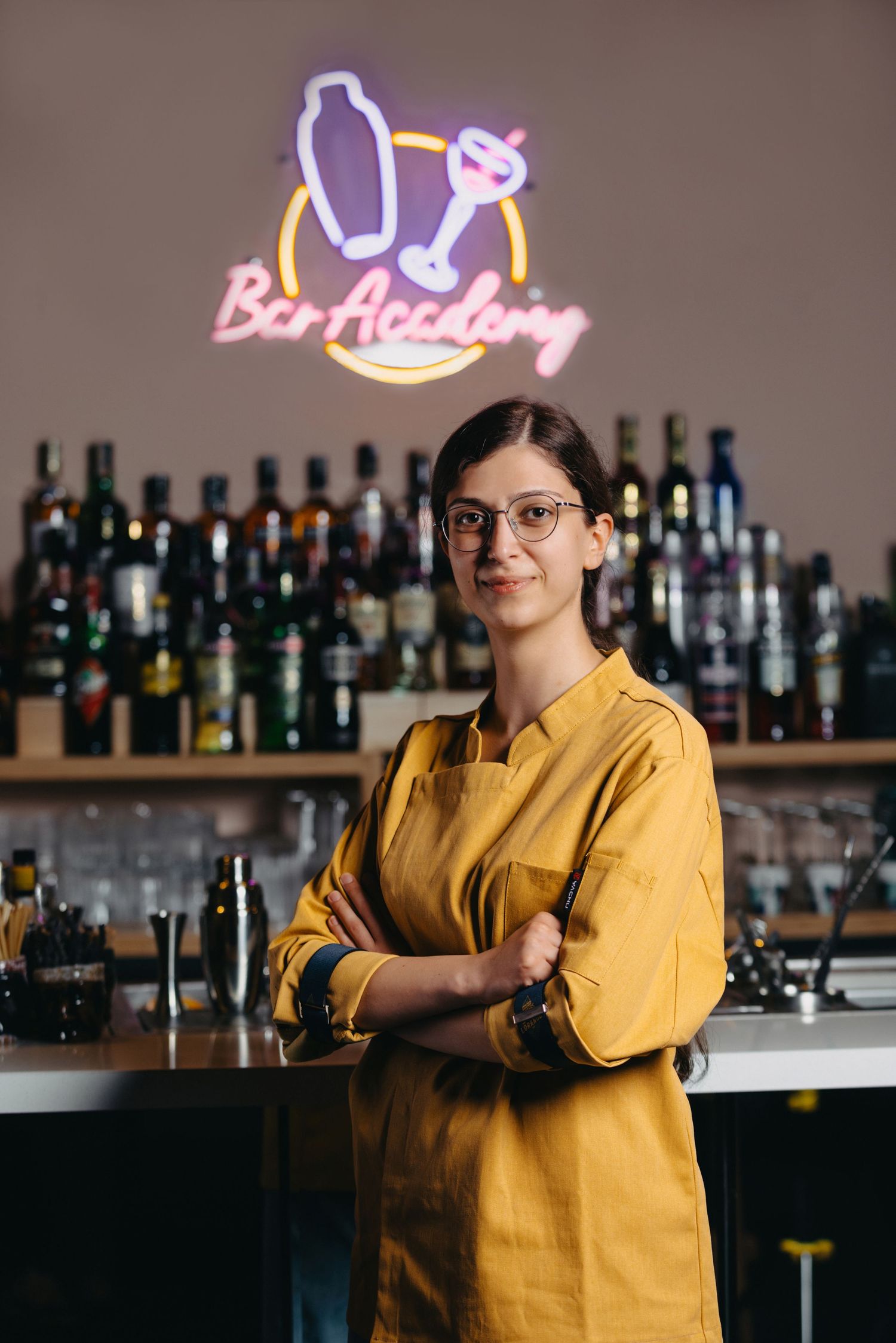 Bartender in yellow shirt poses confidently at bar counter with neon signs and bottles.
