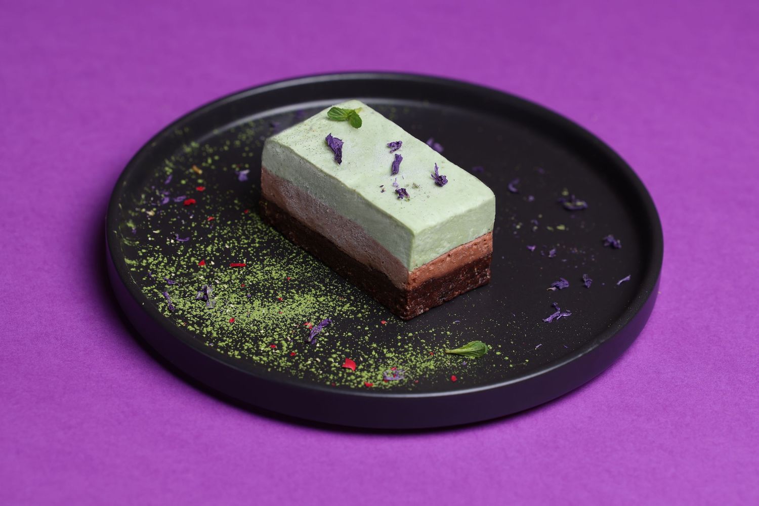 Dessert plating sequence showing a slice of cake with colorful sprinkles on a black plate against purple background.