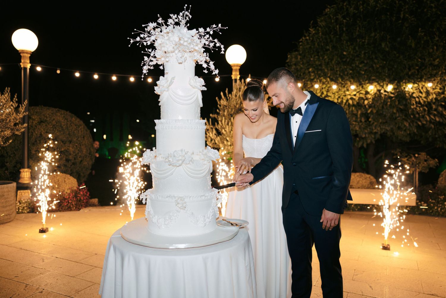 Newlyweds stand beside their wedding cake with sparklers and string lights creating a magical atmosphere.