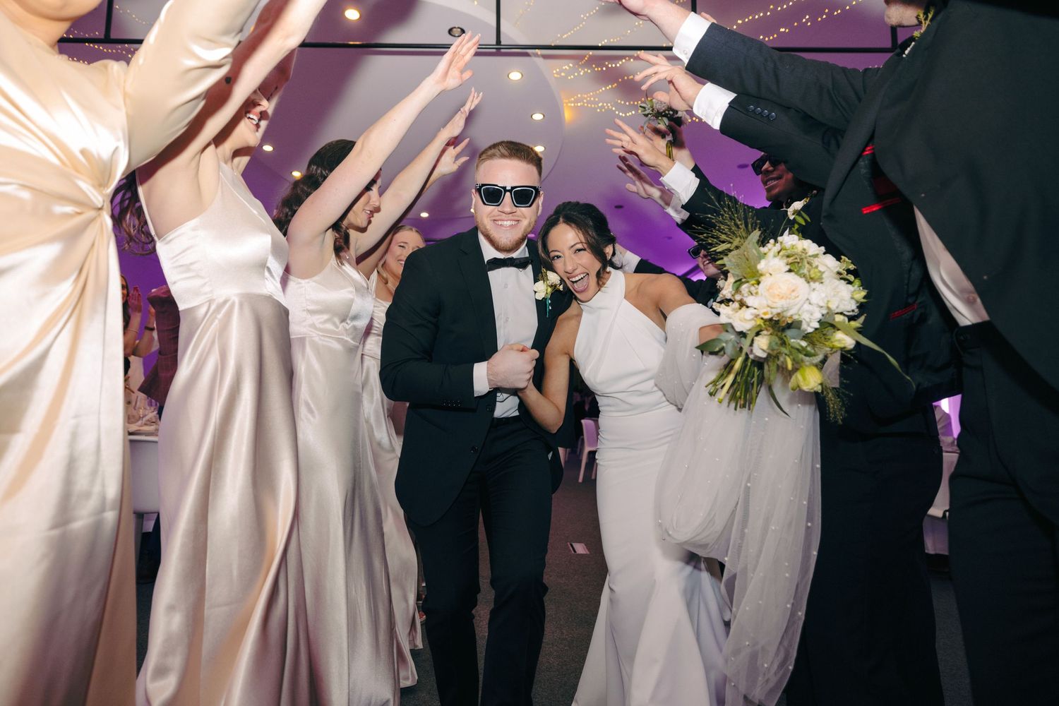 Wedding celebration dance floor with guests in formal attire under purple lighting.