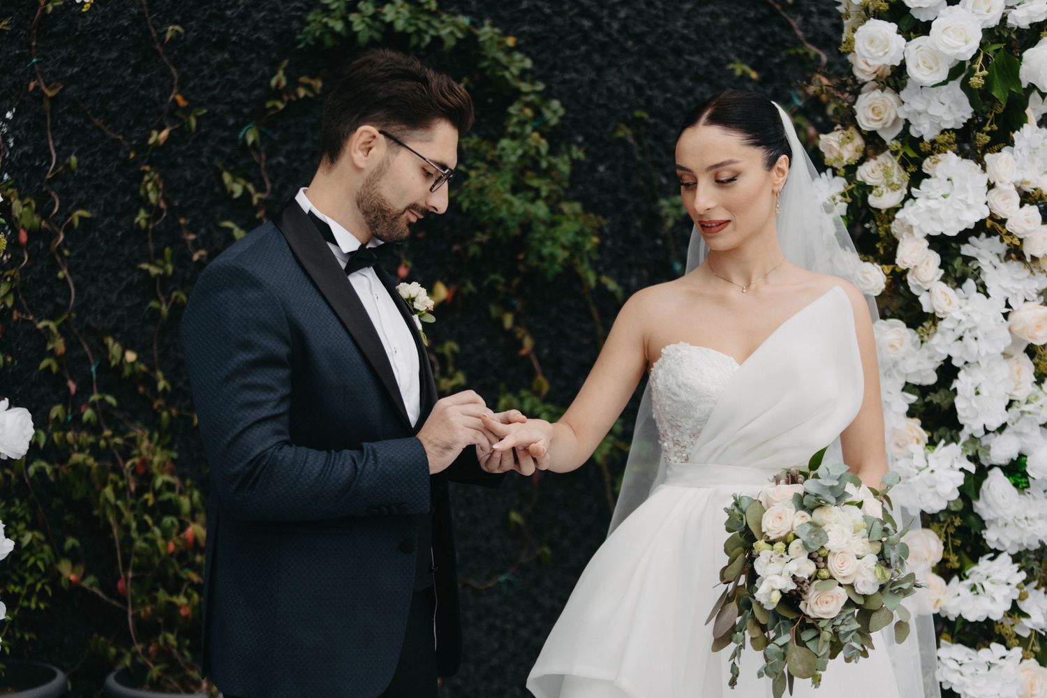 Wedding couple exchanges rings during ceremony against floral wall backdrop.