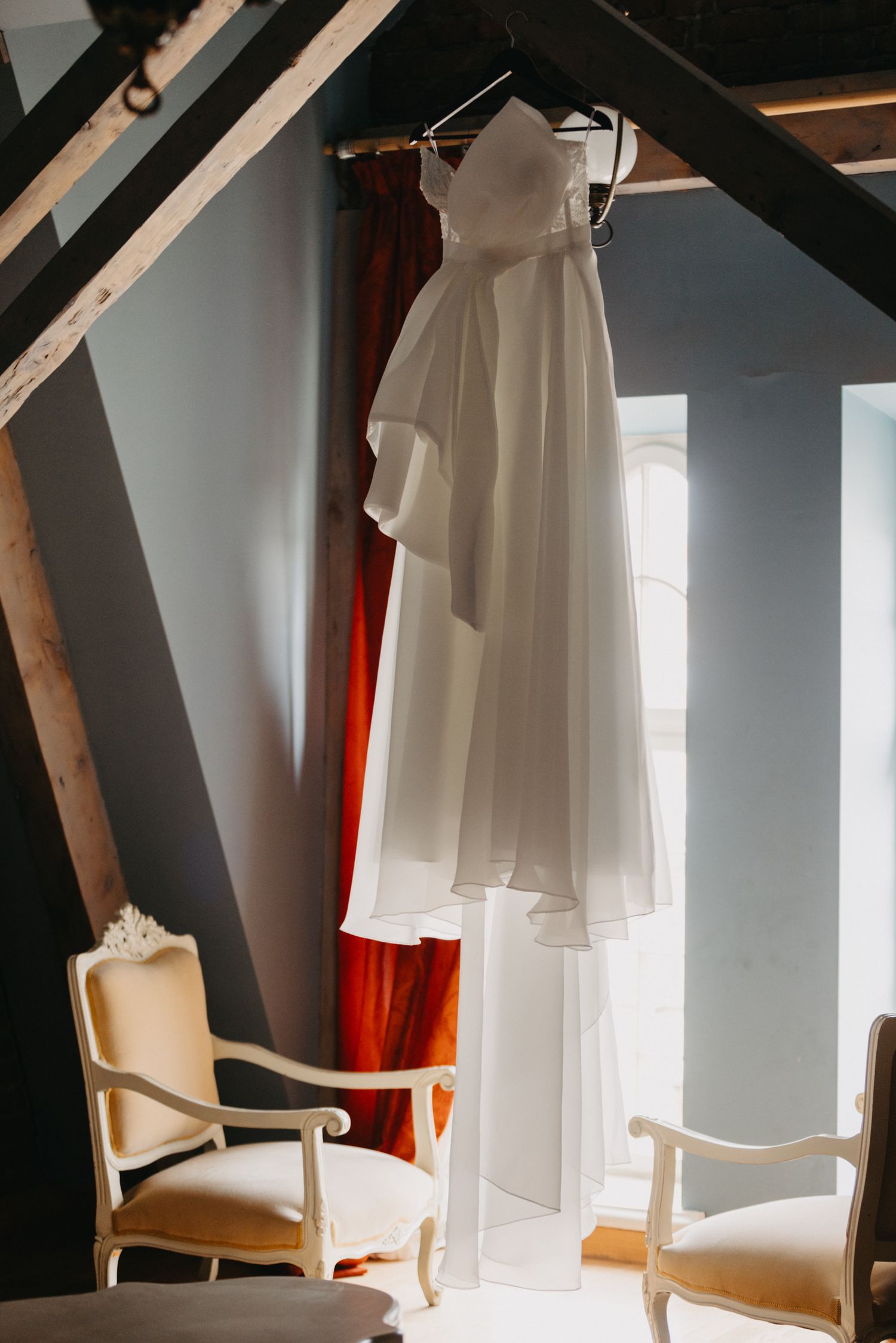 Elegant white wedding dress hanging in a room with cream-colored chairs and dramatic black beams.