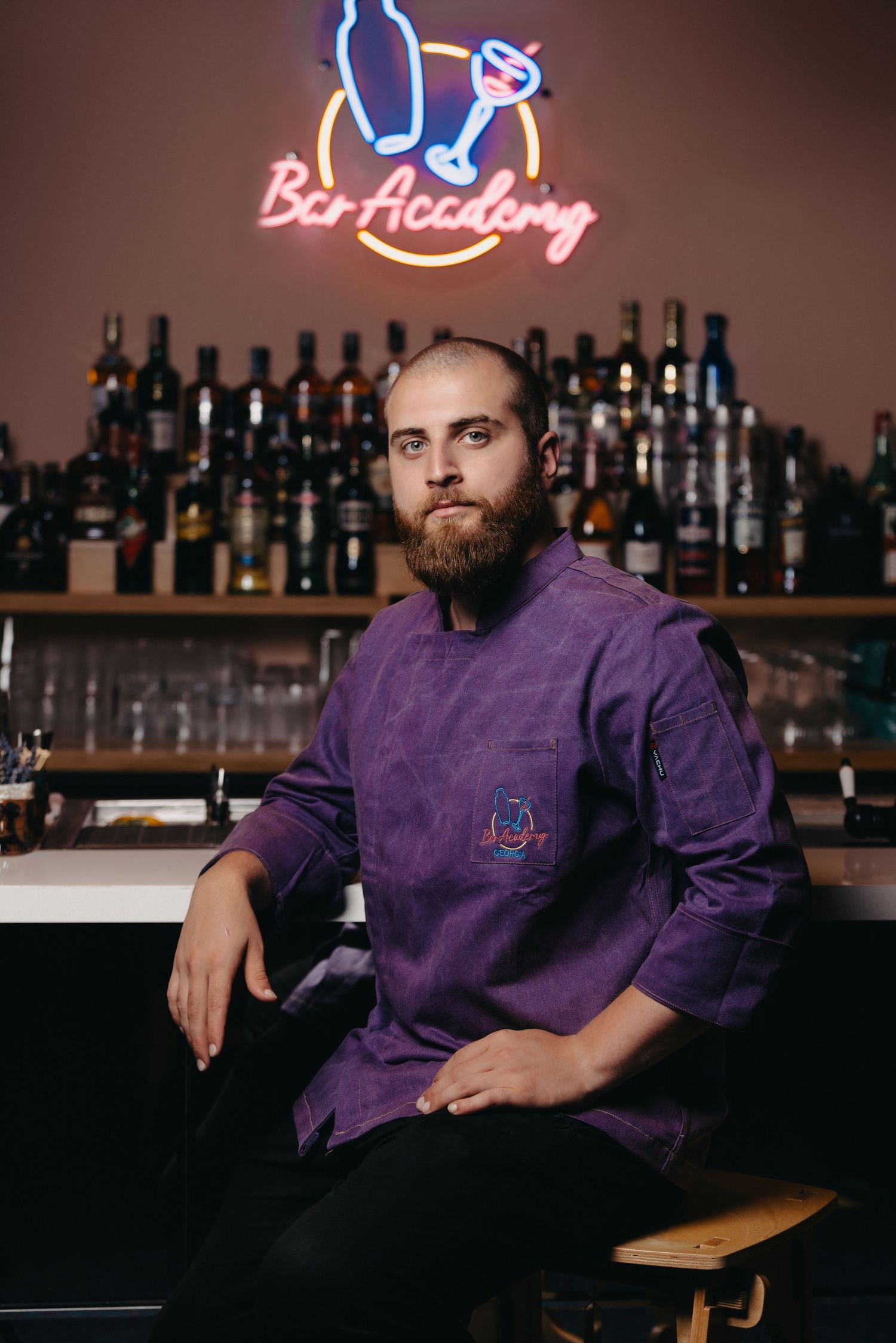 Professional bartender in purple uniform posing at bar counter with bottles in background.
