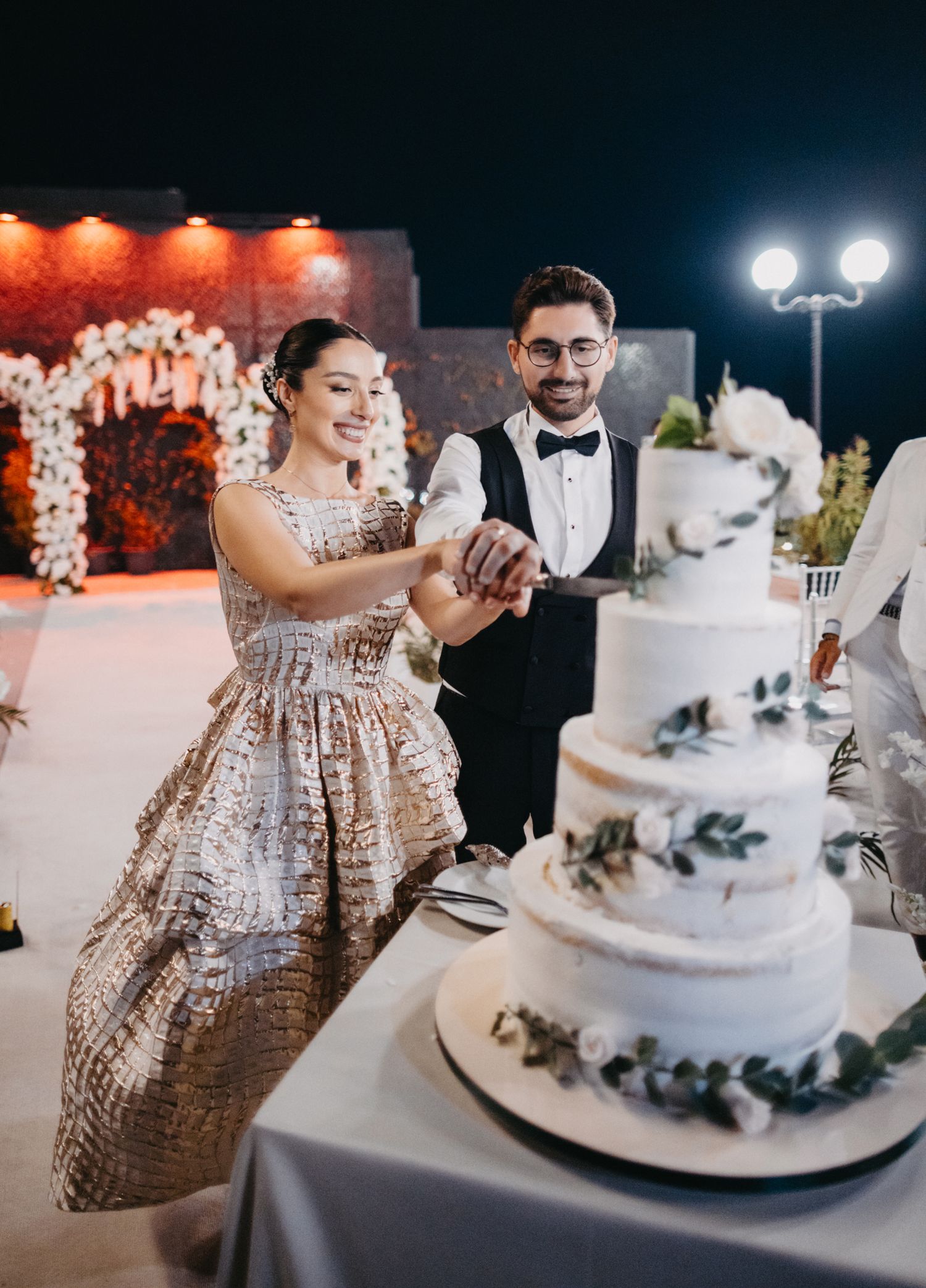 Couple cuts their elegant white wedding cake decorated with greenery at evening reception.