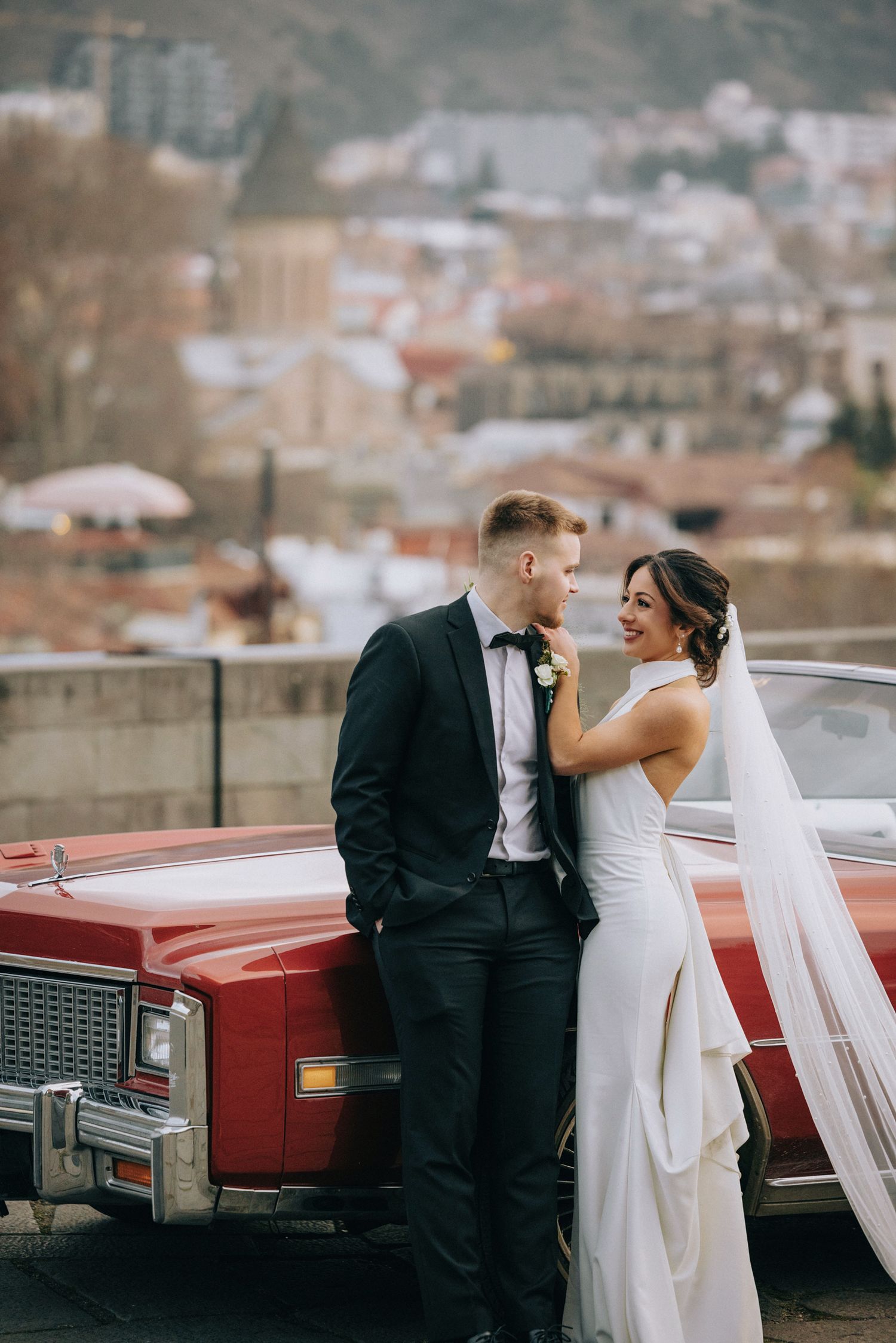 Wedding couple poses by vintage red car with city backdrop.
