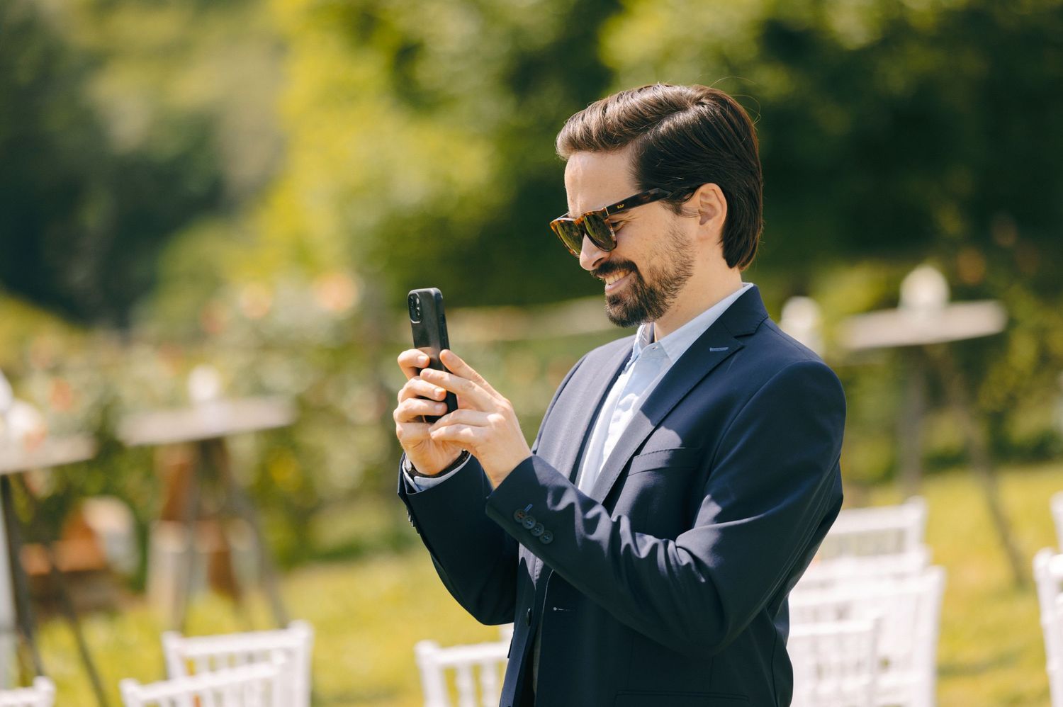 A wedding guest in a navy suit checks their phone during an outdoor ceremony.