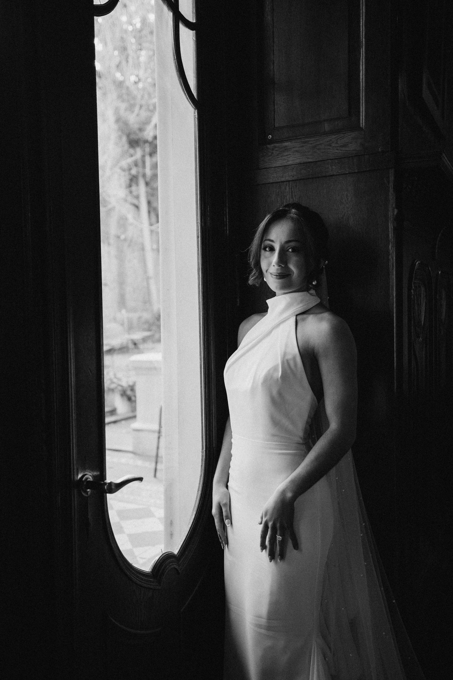 Dramatic black and white photo of a bride in a sleek white dress standing in a doorway.