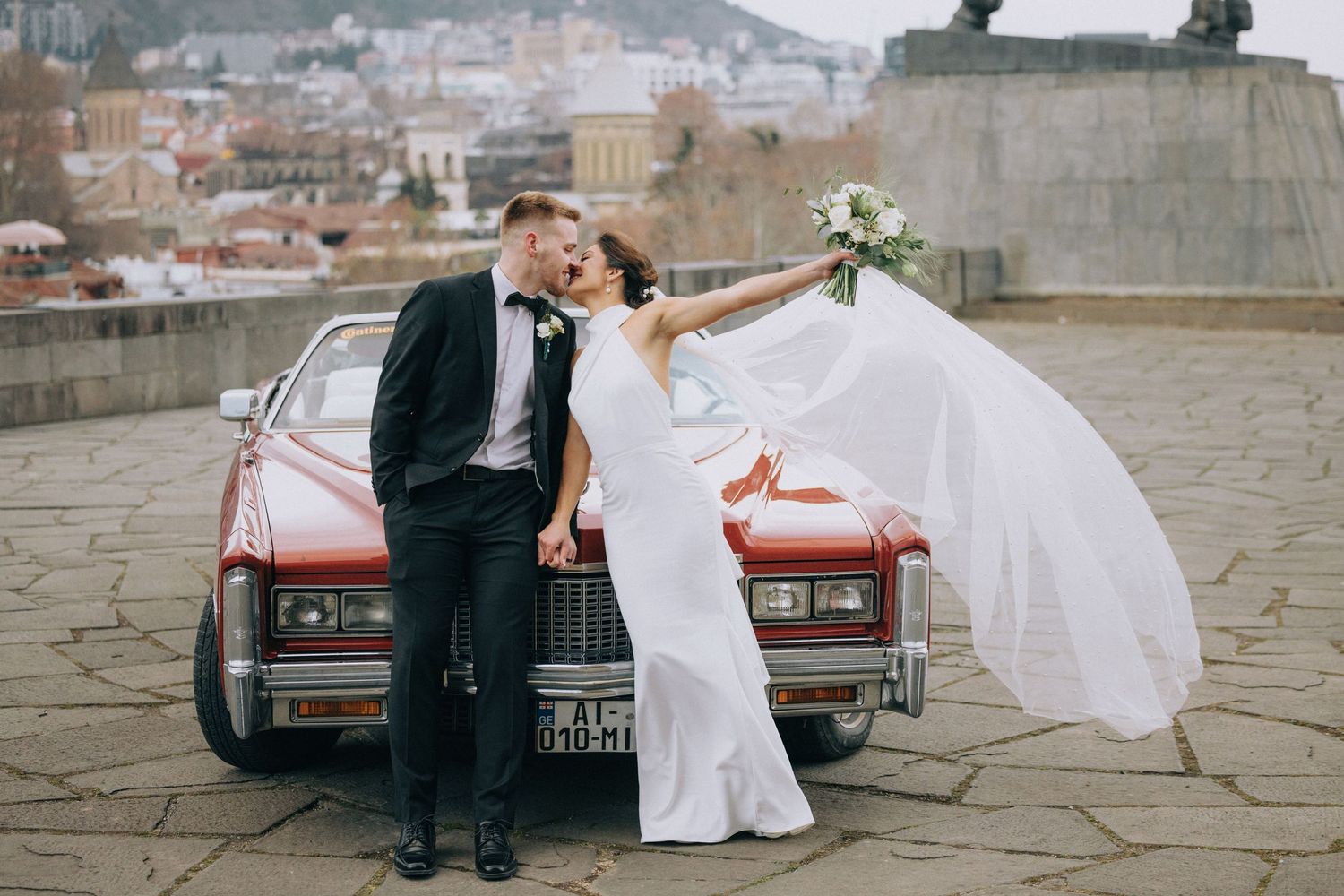 Elegant wedding scene with classic red car on stone pavement.