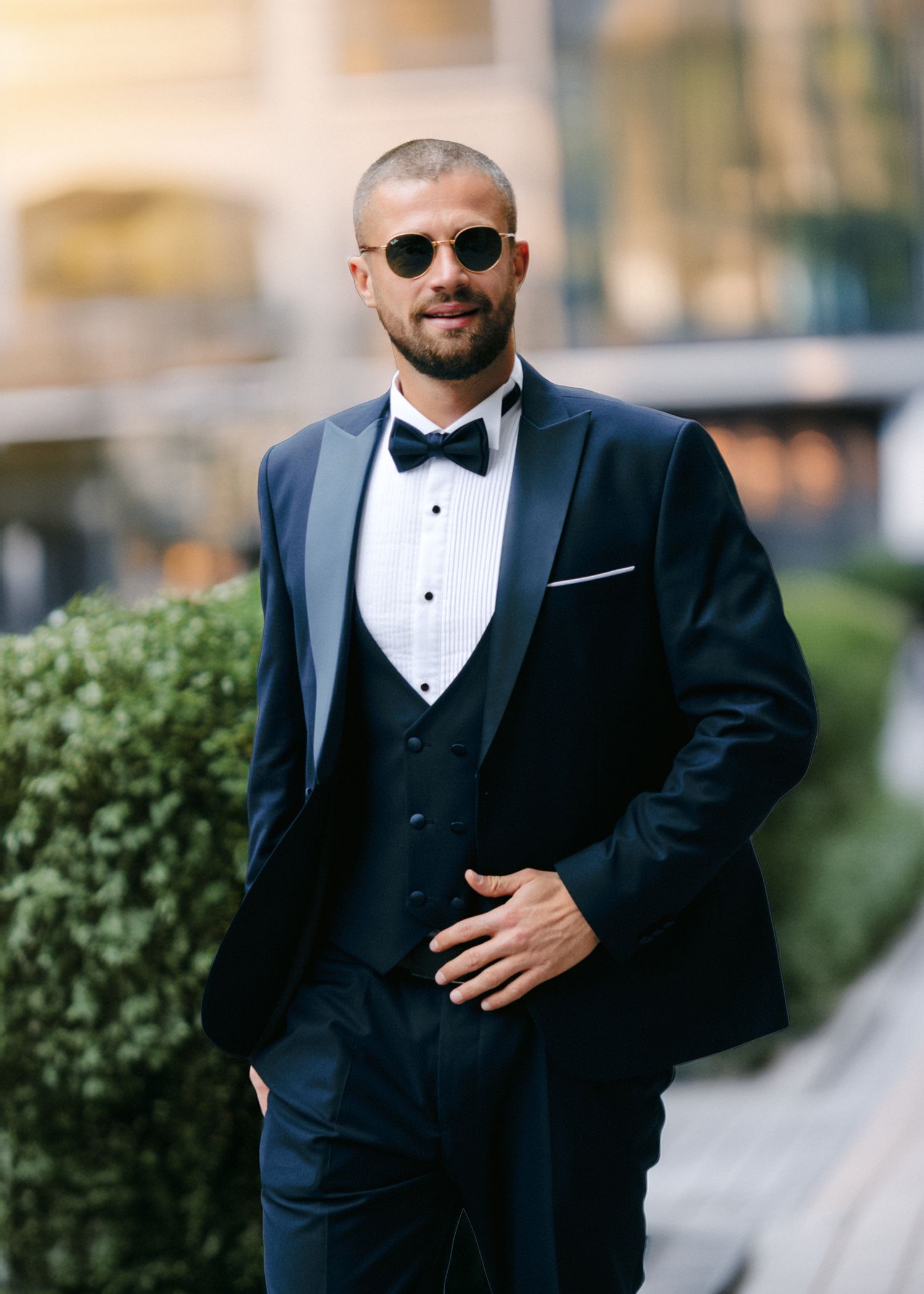 Groom in classic black tuxedo and bow tie poses confidently on a sidewalk.
