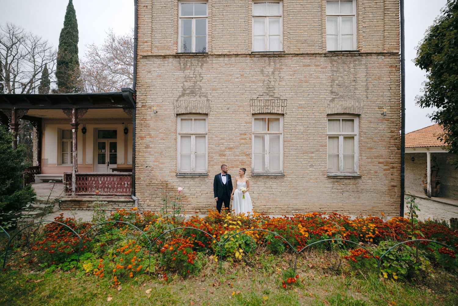 Wedding couple photographed in front of vintage brick building with blooming garden.