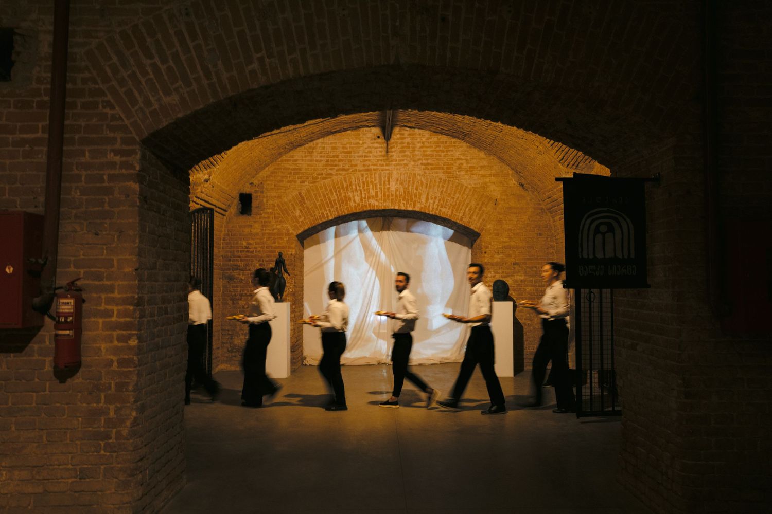 Dark tunnel corridor with brick archways illuminated by warm lighting and walking figures in shadow.