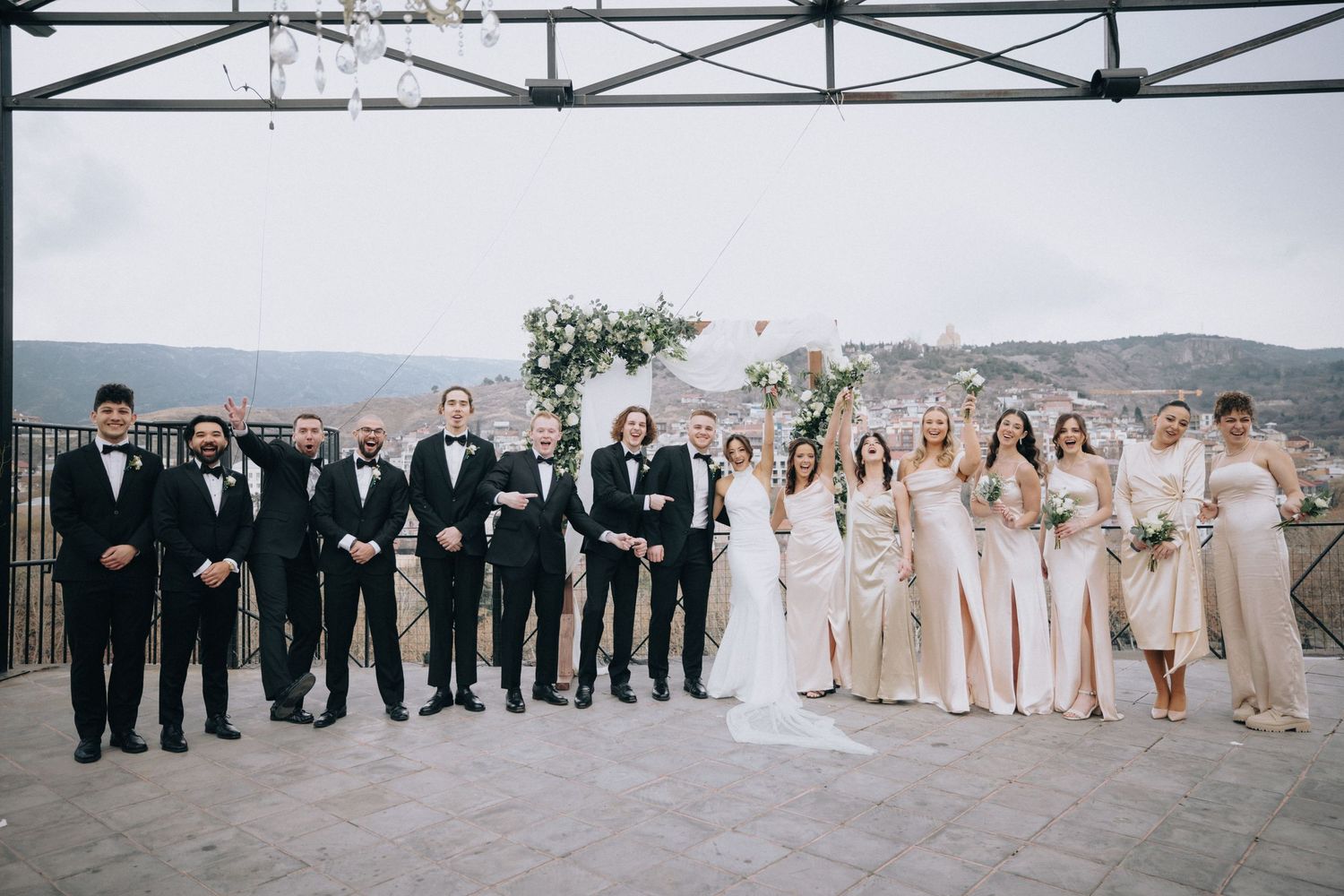 Wedding party in black and beige attire poses together on outdoor terrace with mountain backdrop.
