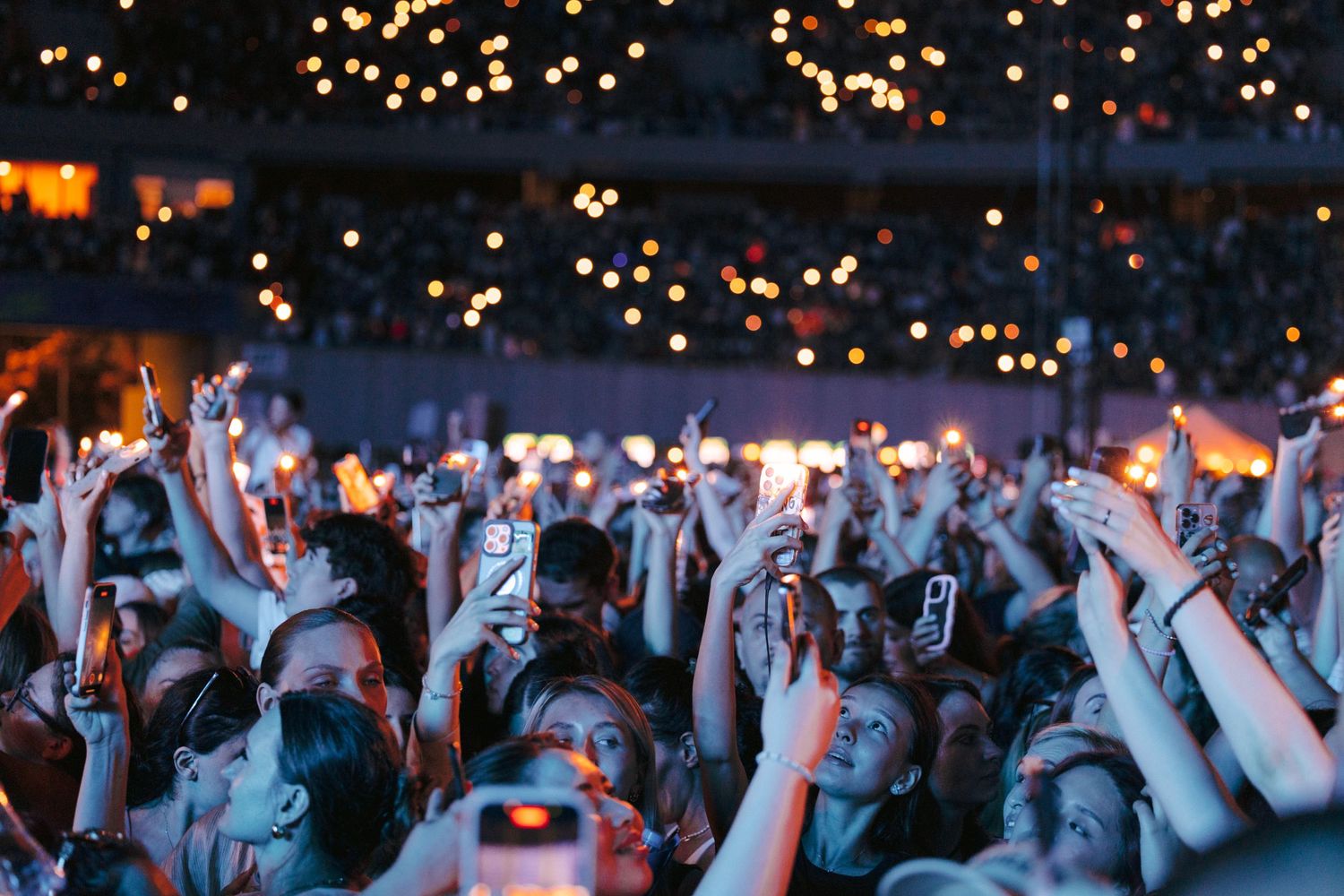 Enthusiastic concert crowd raises hands with phone lights glowing in stadium atmosphere.