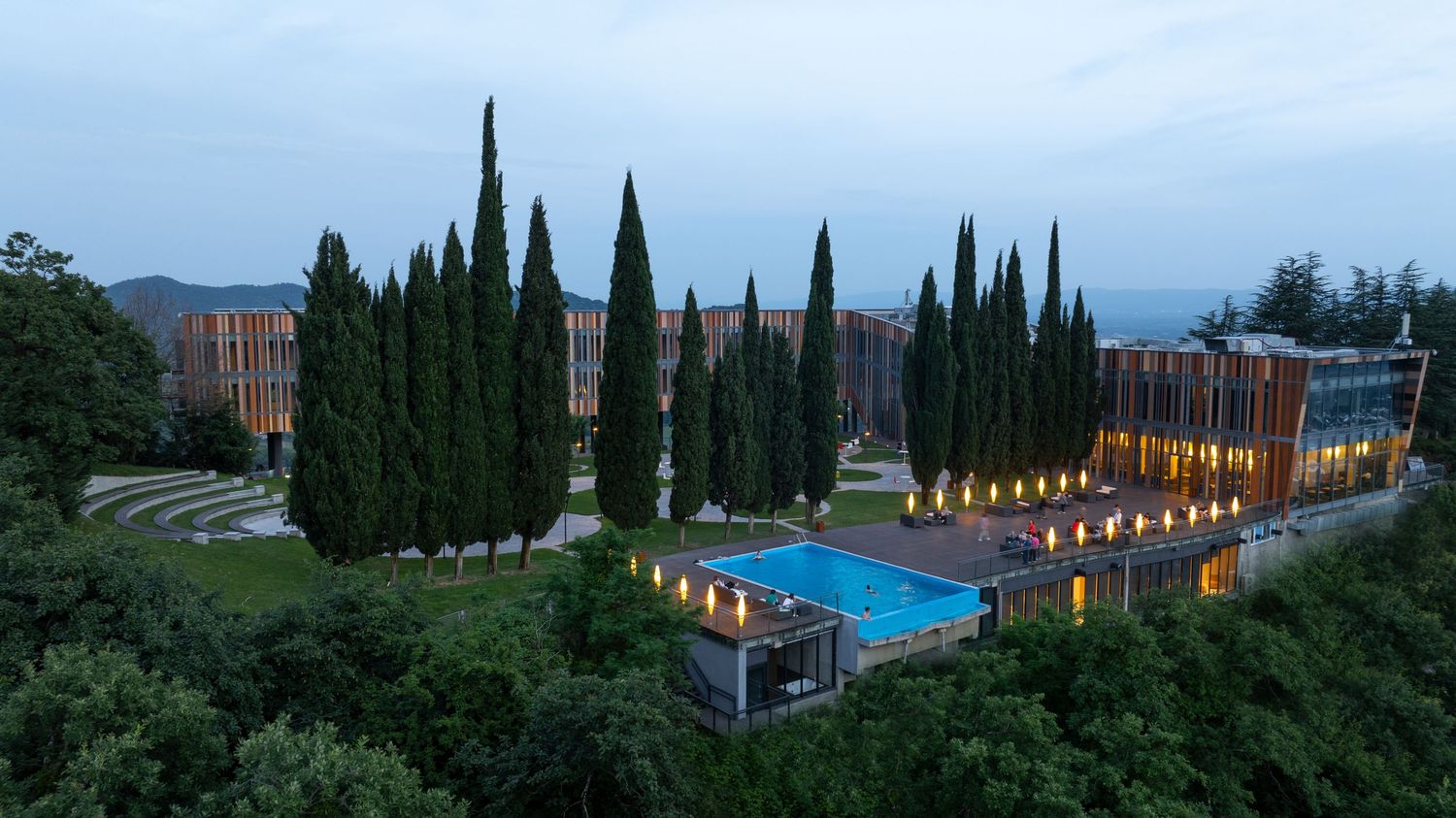 Aerial panoramic view of a modern luxury resort with swimming pool and cypress trees at dusk.