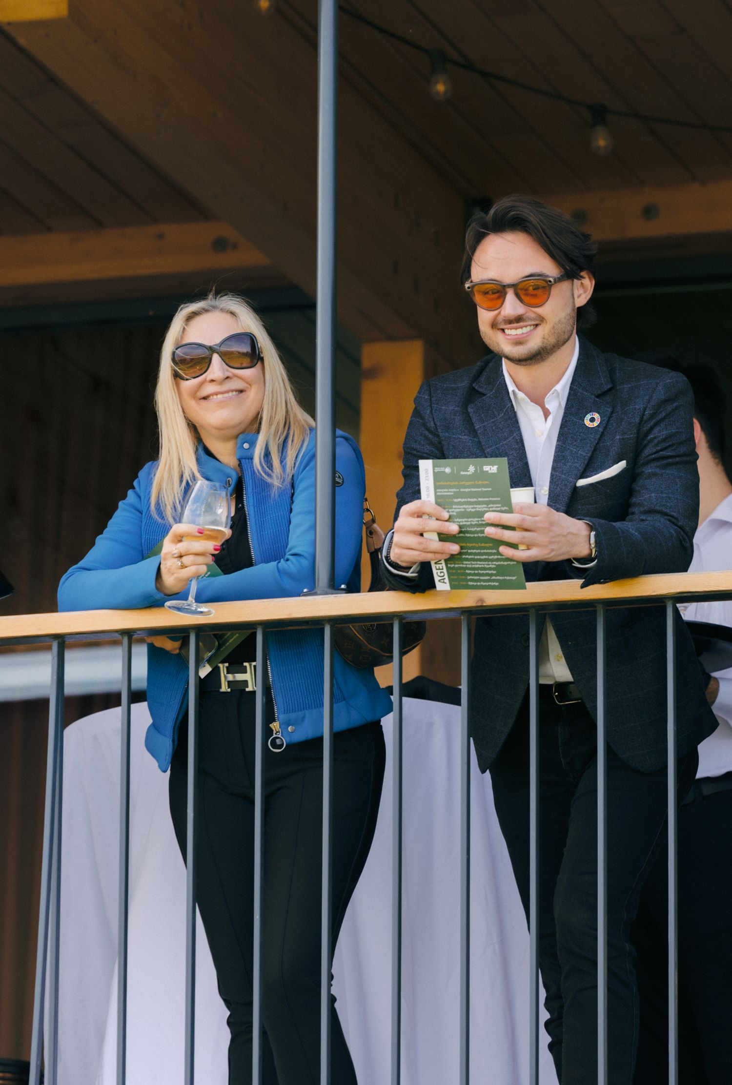 Attendees in blue shirts and sunglasses enjoying drinks at an outdoor event balcony.