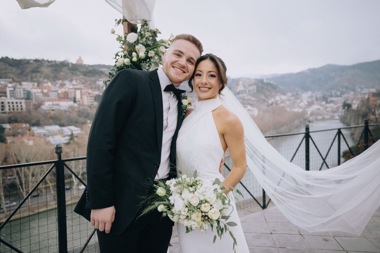 Newlywed couple poses together on mountain terrace with flowing veil and white floral bouquet.