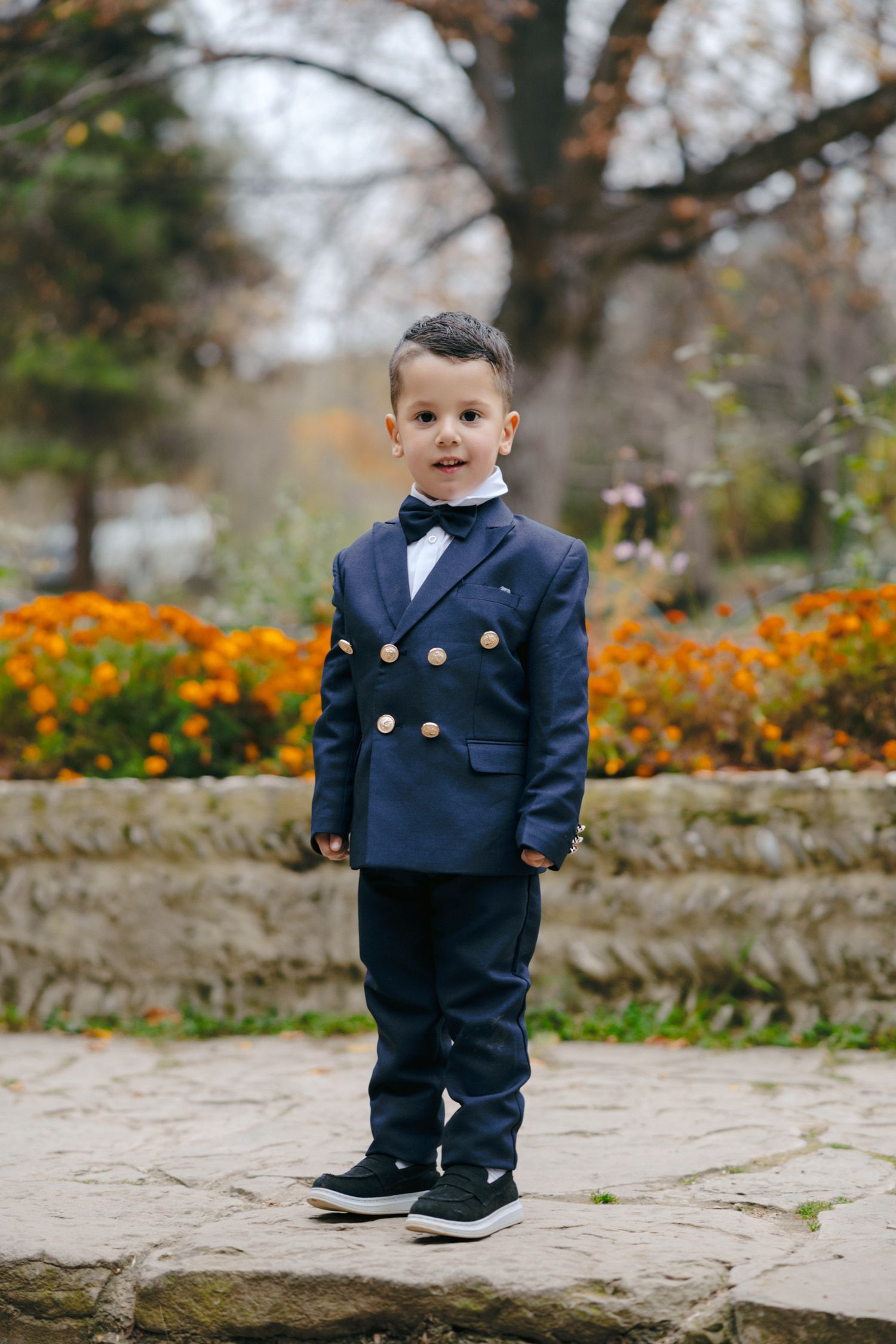 Young ring bearer in navy dress uniform stands on stone platform near orange flowers.