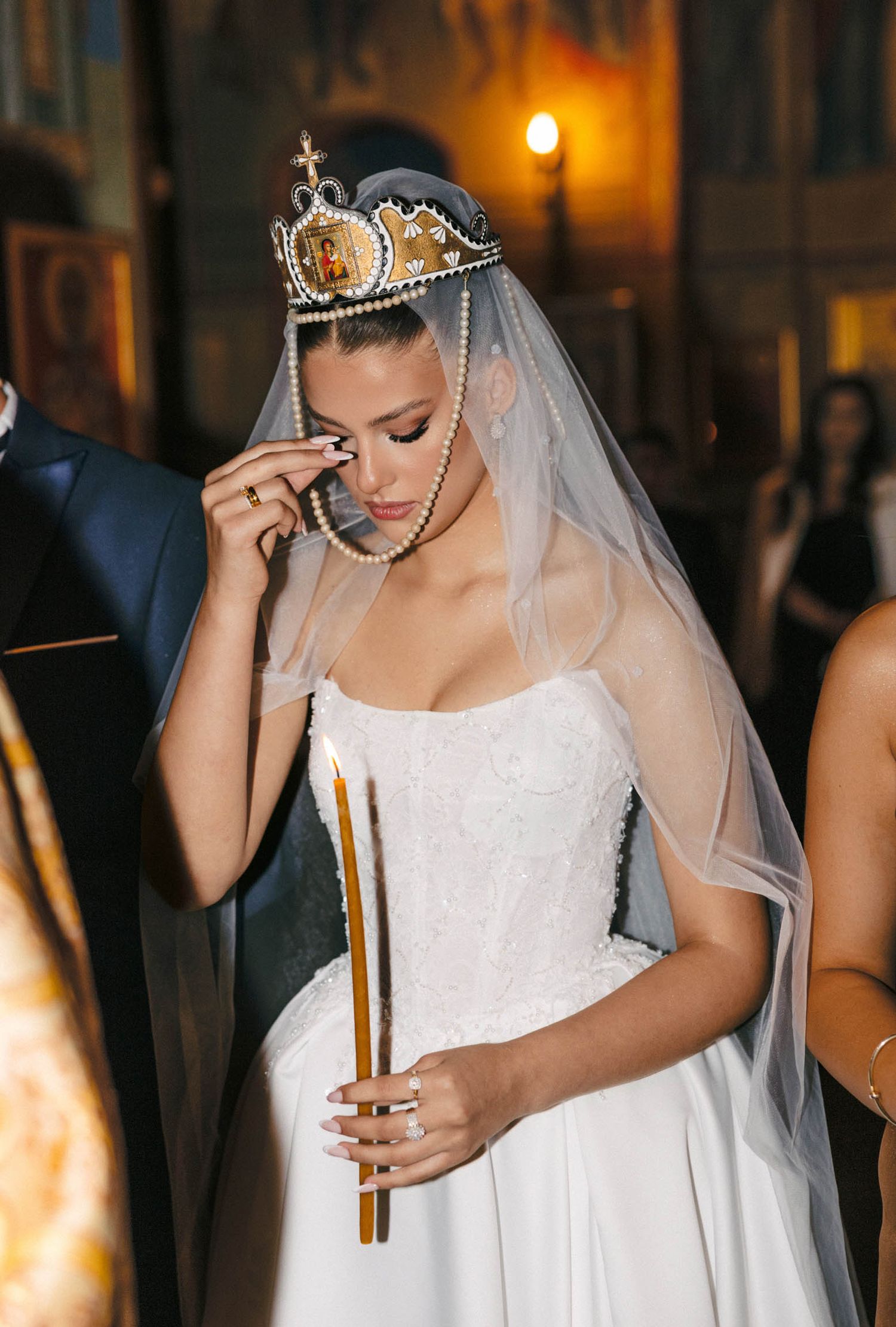 Bride in white dress and crown veil holding a ceremonial candle during evening ceremony.