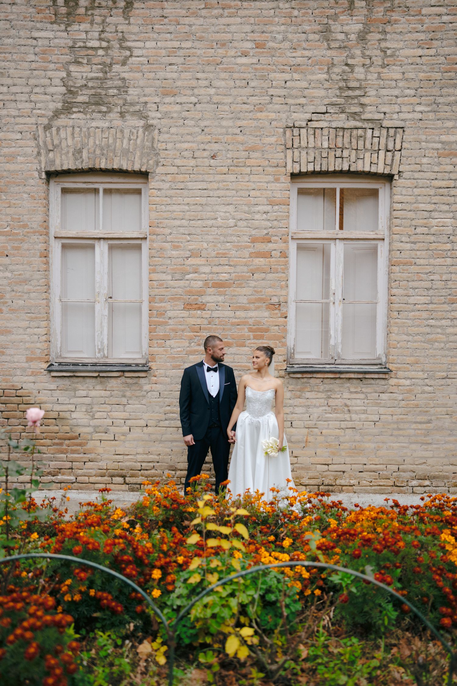 Elegant couple stands among orange flowers against historic brick building with tall windows.