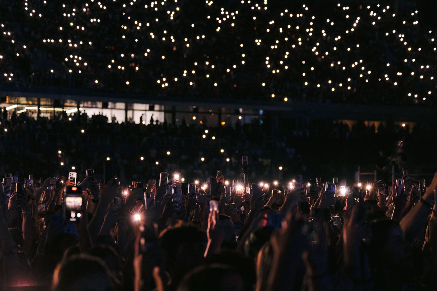 Concert crowd holds up phone lights creating a magical starlit atmosphere in darkness.