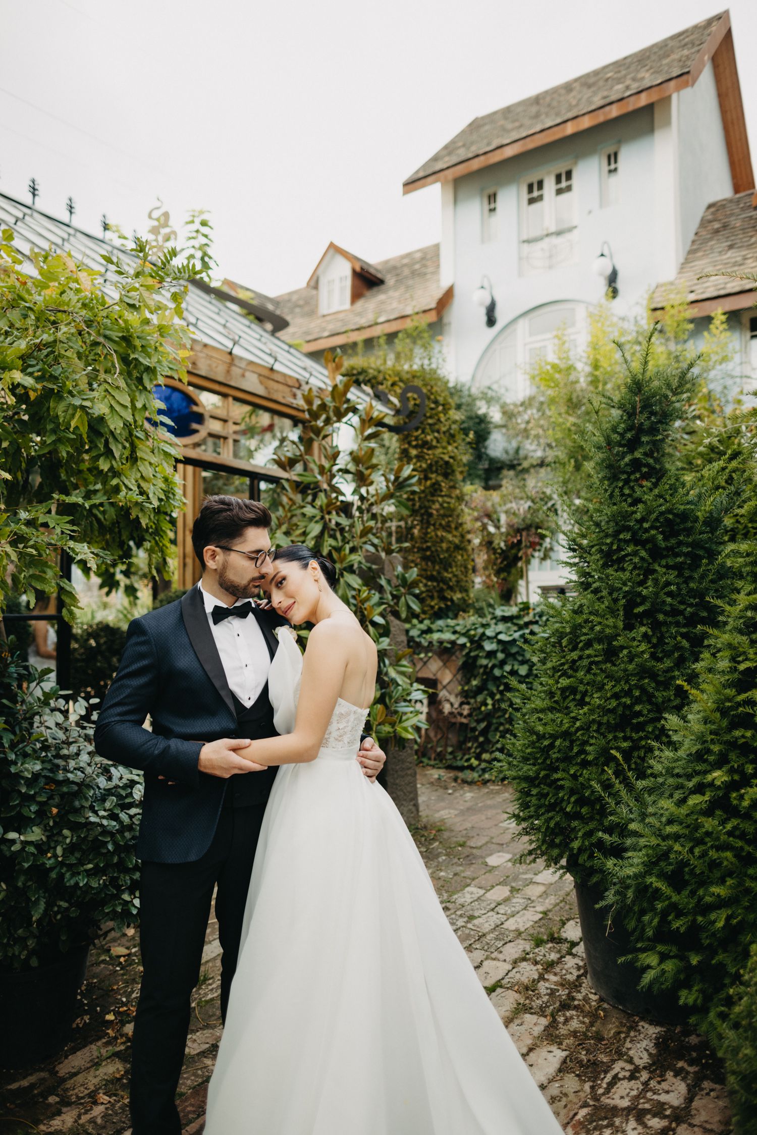 Romantic wedding couple shares intimate moment in a charming garden with rustic architecture.