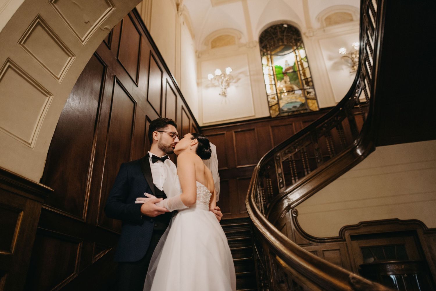 Wedding couple shares intimate moment on grand wooden staircase in historic venue.