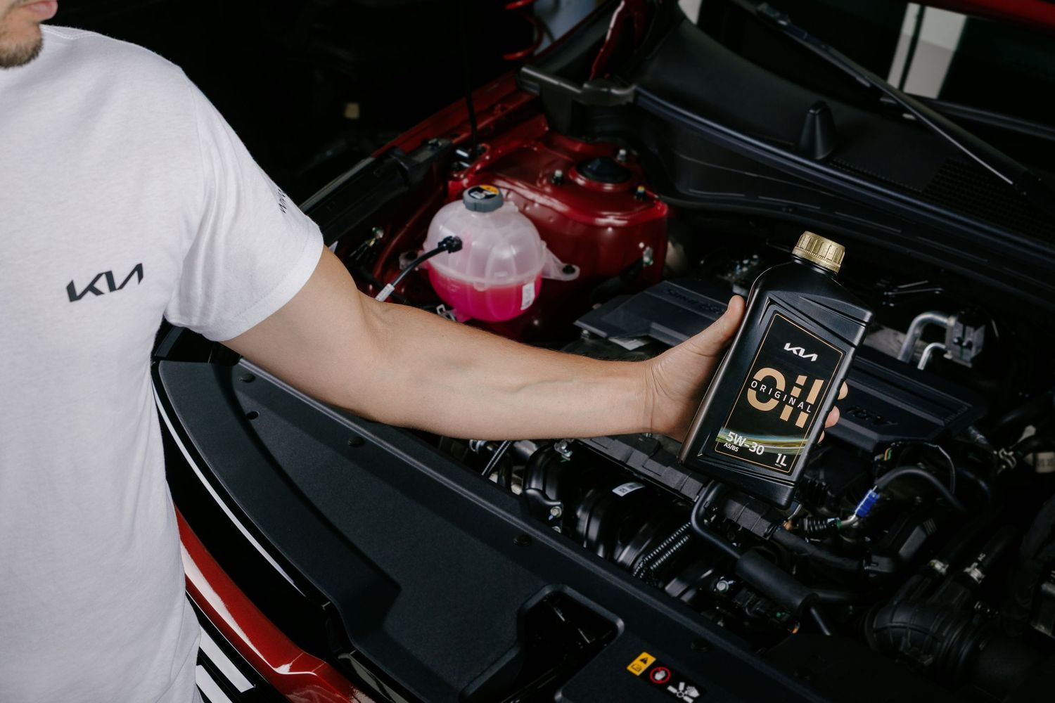 A Kia mechanic holds a bottle of motor oil near an open red car engine hood during a routine maintenance service.
