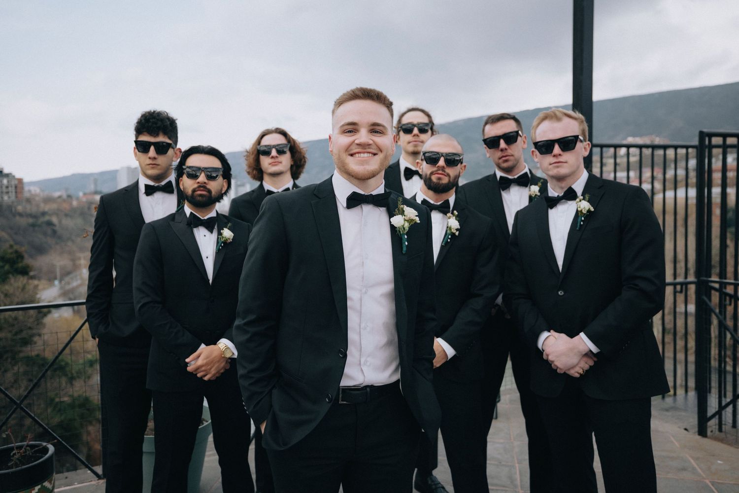 Groomsmen in black tuxedos and sunglasses pose together on outdoor terrace.