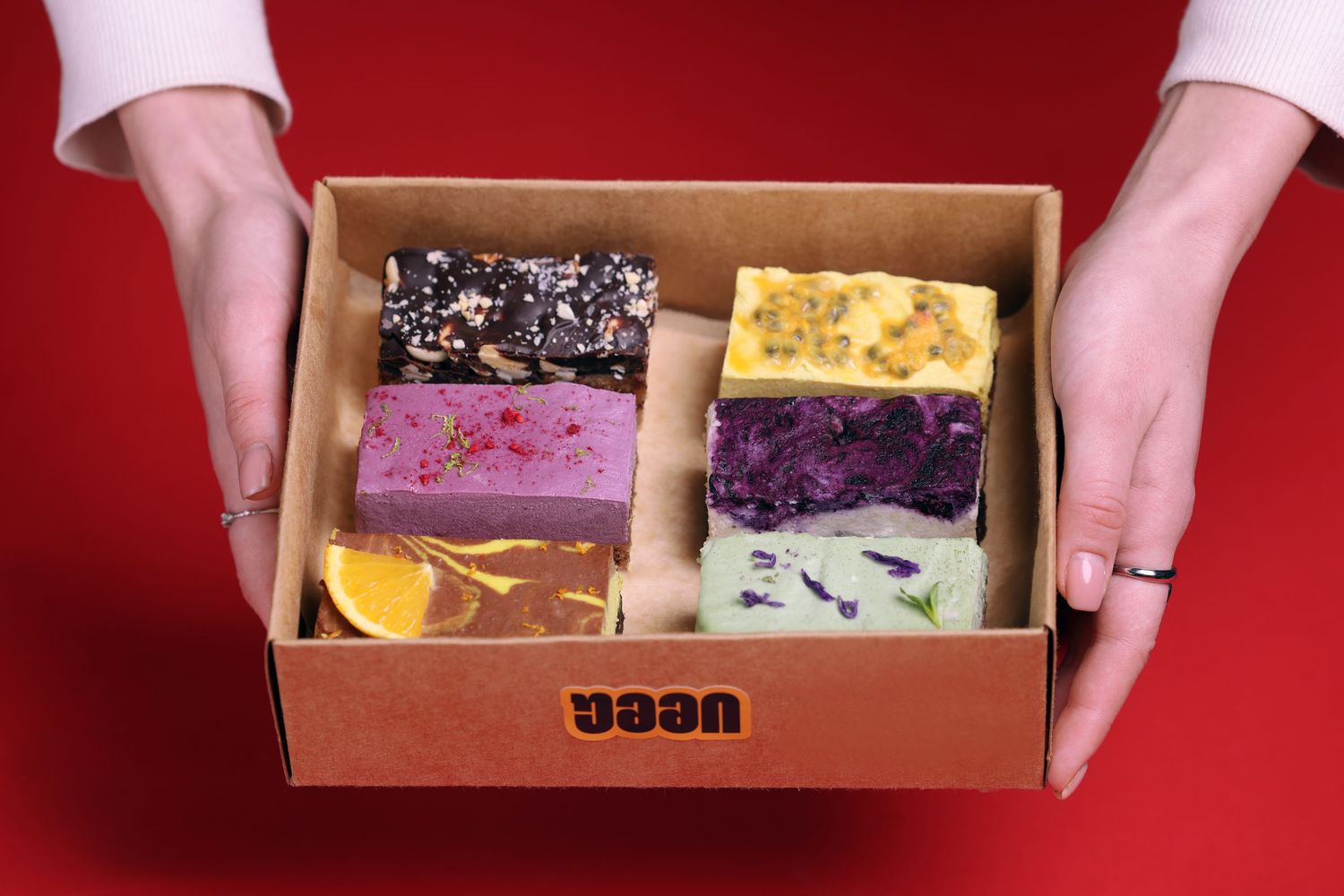 Hands holding dessert box with colorful pastries and treats against red background.