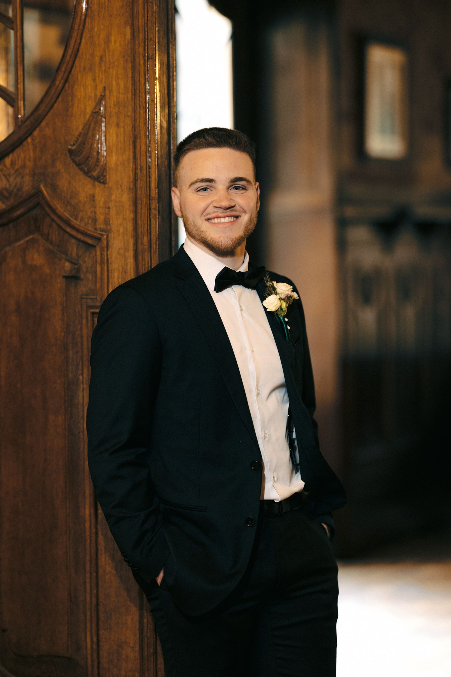 Groom in classic black tuxedo and bow tie poses against wooden paneling.