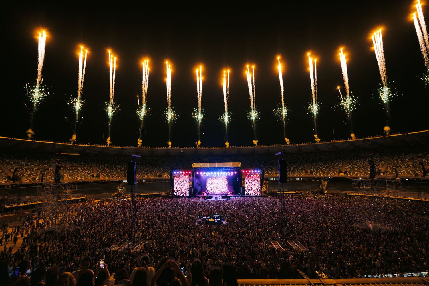Stadium concert with pyrotechnic display creating golden lights above massive crowd.