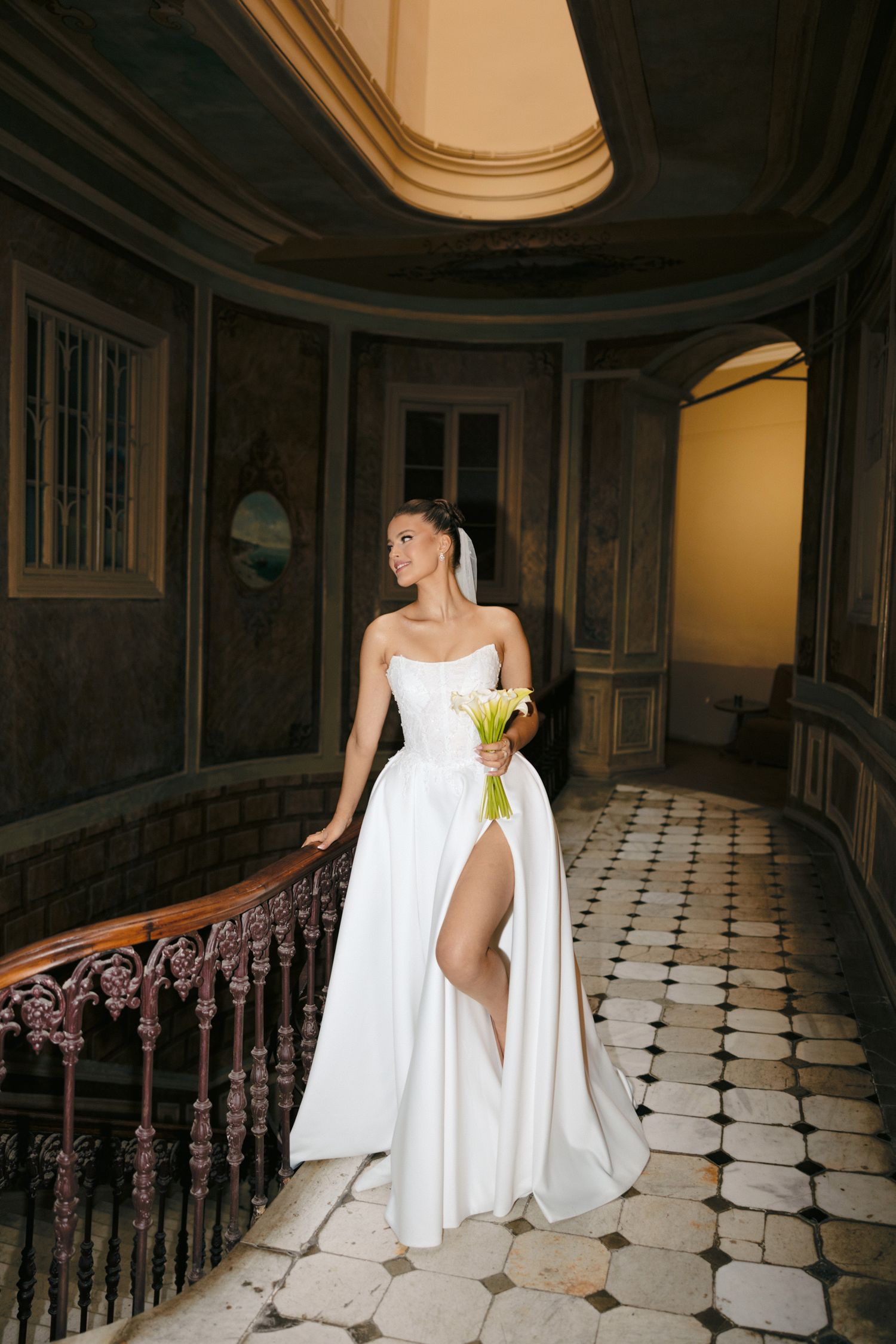 Bride in elegant white gown with thigh-high slit poses in historic building interior.